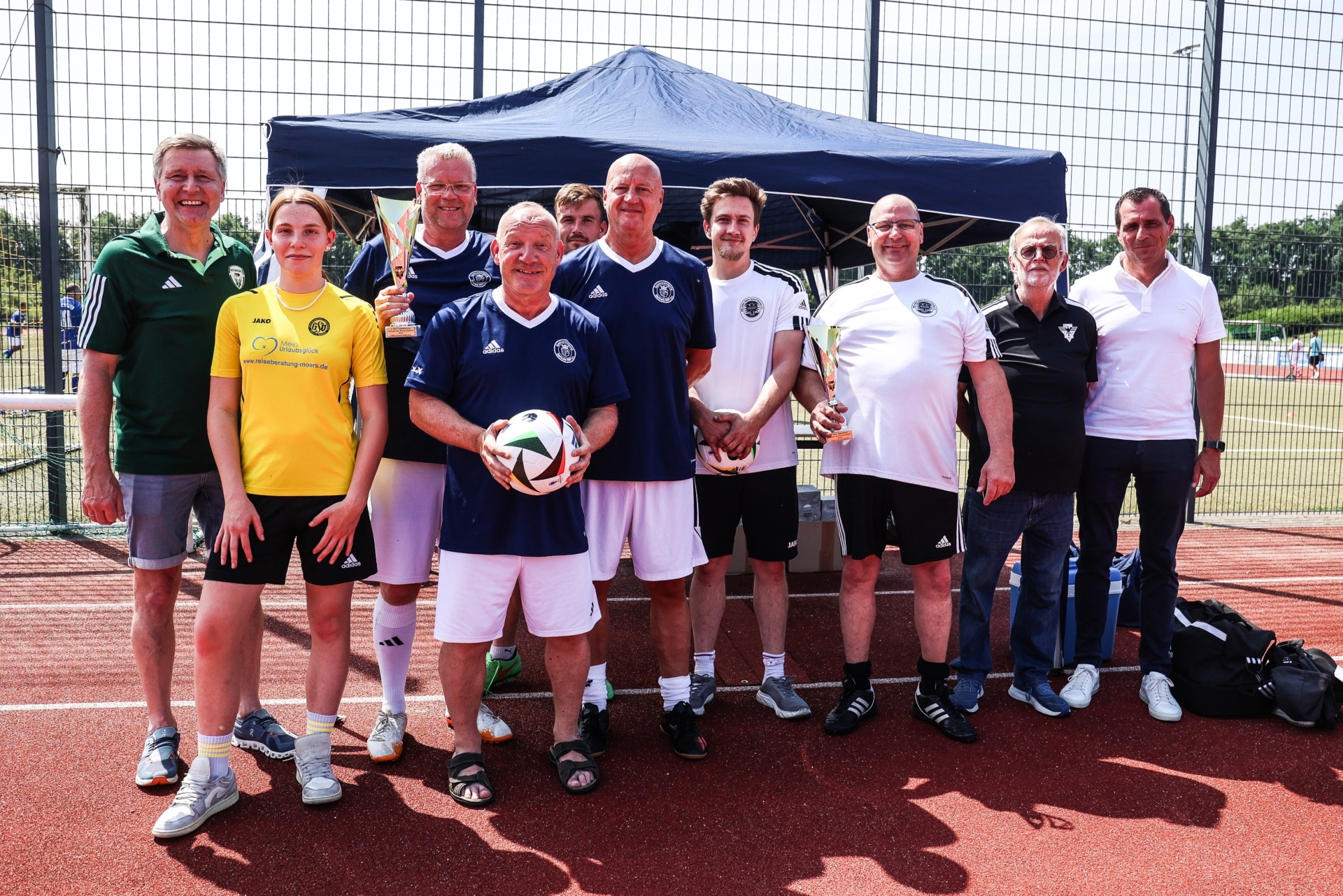 [{"clang_id":1,"value":"Gruppe von neun Personen steht auf einem roten Sportplatz vor einem blauen Zelt. Die meisten tragen Sportkleidung, einige halten einen Fußball."}]