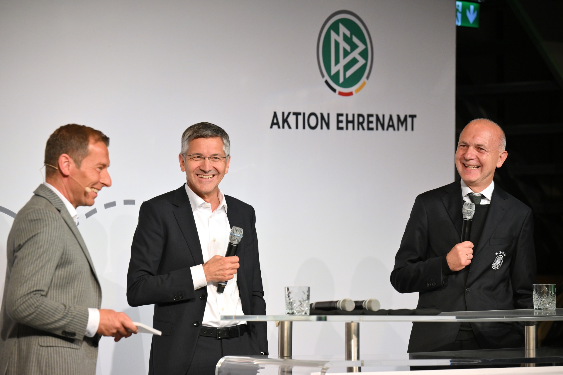 [{"clang_id":1,"value":"Drei Männer in Anzügen stehen an einem Rednerpult vor einer weißen Wand mit grünem Logo und dem Text \"AKTION EHRENAMT\". Alle drei lächeln."}]