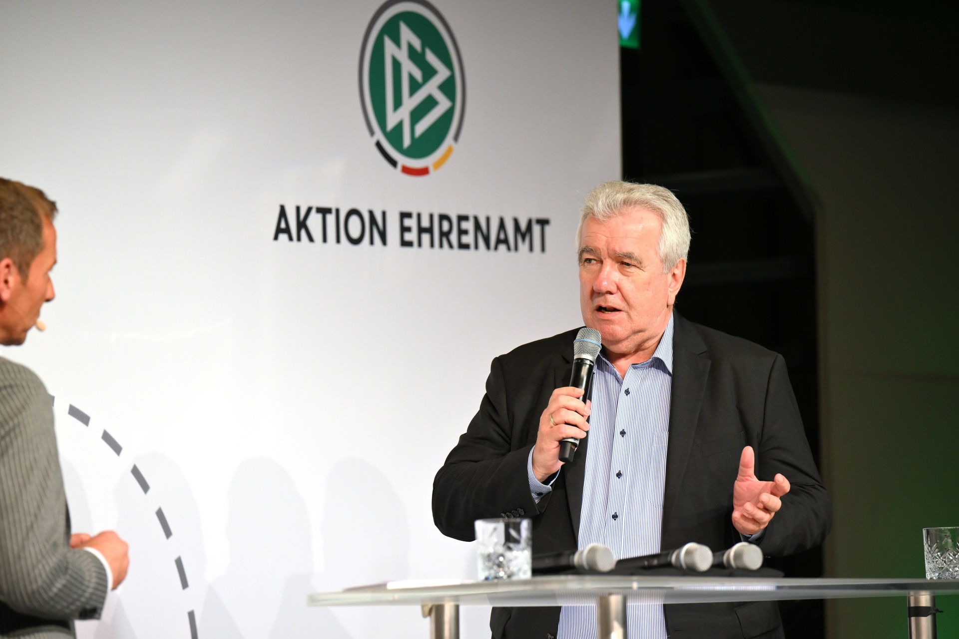 [{"clang_id":1,"value":"Zwei Männer an einem Tisch vor einem Banner mit dem Logo \"AKTION EHRENAMT\". Der Mann rechts spricht in ein Mikrofon, der andere hört zu."}]