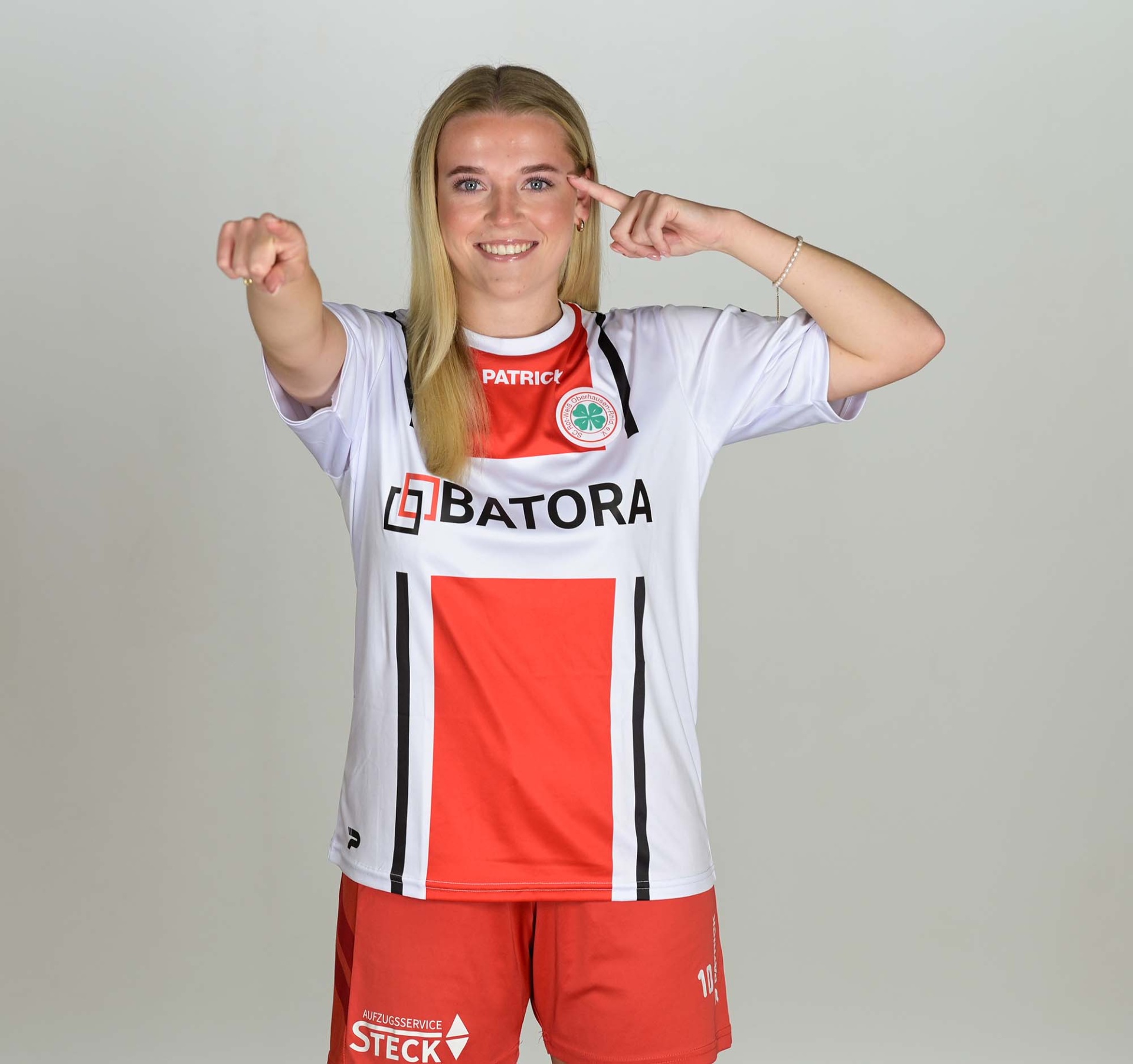 [{"clang_id":1,"value":"Frau in rotem und weißem Fußballtrikot mit Nummer 10 und Shorts, macht mit Fingern das \"Schuss\"-Signal vor grauem Hintergrund."}]