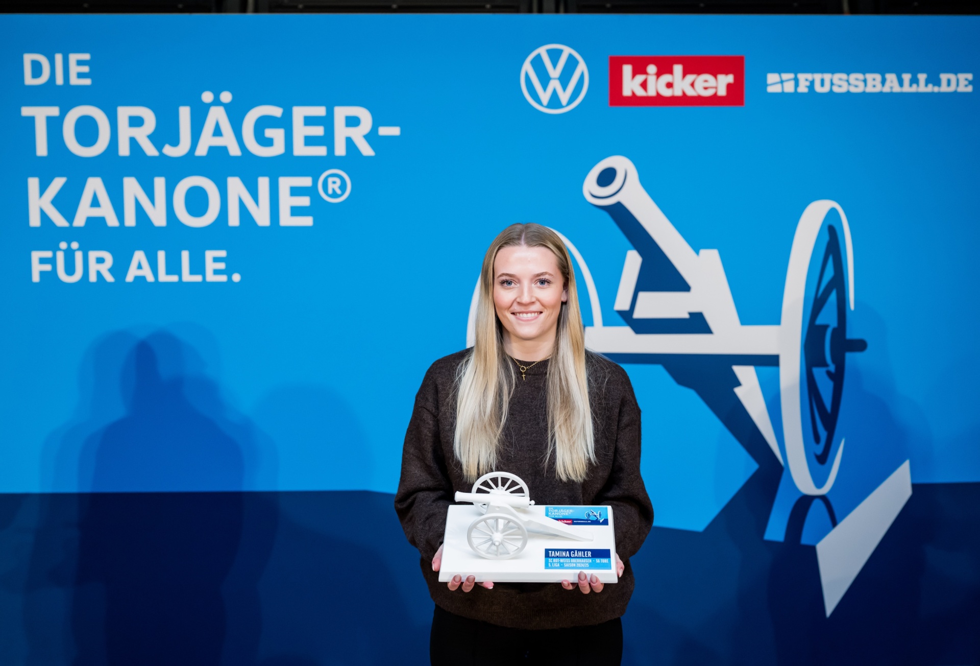 [{"clang_id":1,"value":"Frau mit Torjägerkanone. Sie steht vor einem blauen Hintergrund mit Logos von Volkswagen, Kicker und Fussball.de. Die Kanone ist silber und tropfenförmig."}]