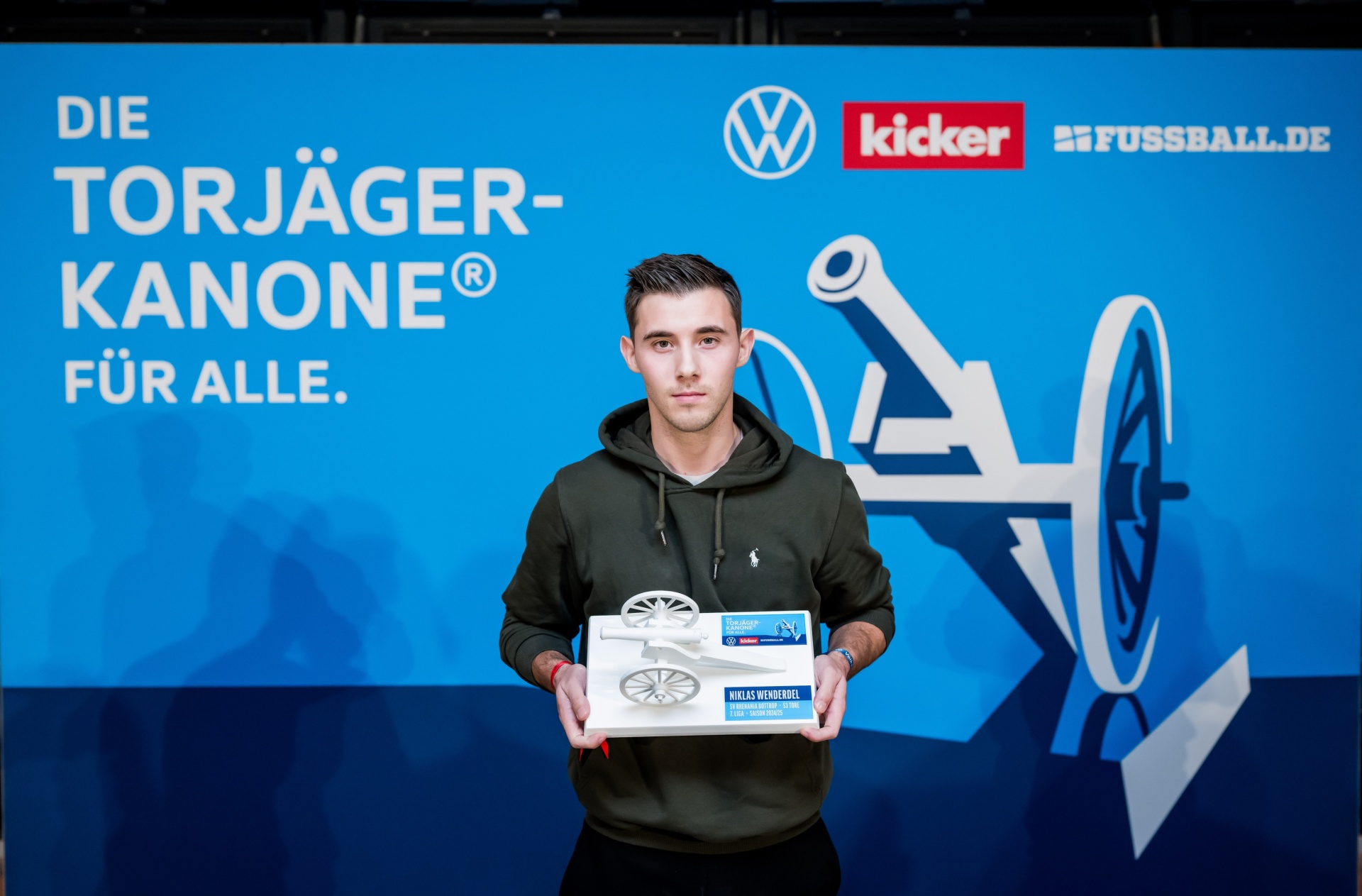[{"clang_id":1,"value":"Junger Mann mit dunkler Kleidung hält eine silberne Trophäe und eine kleine blaue Box. Hintergrund ist dunkelblau mit Logos und Text zur \"Torjägerkanone\"."}]