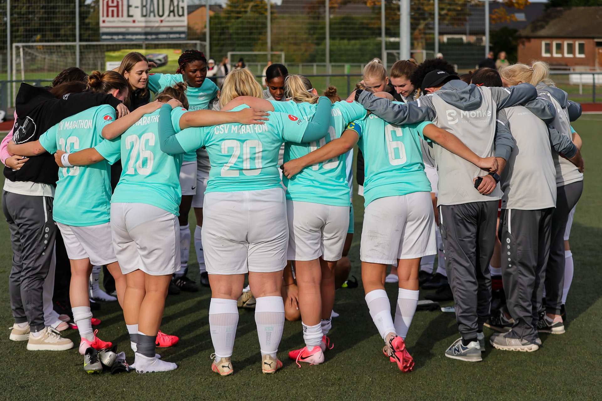 [{"clang_id":1,"value":"Frauenfußballteam im Kreis auf grünem Rasen, Arme um Schultern. Spieler tragen türkisfarbene Trikots mit Nummern, weiße Shorts und Socken. Im Hintergrund eine Tribüne und Werbebanner."}]