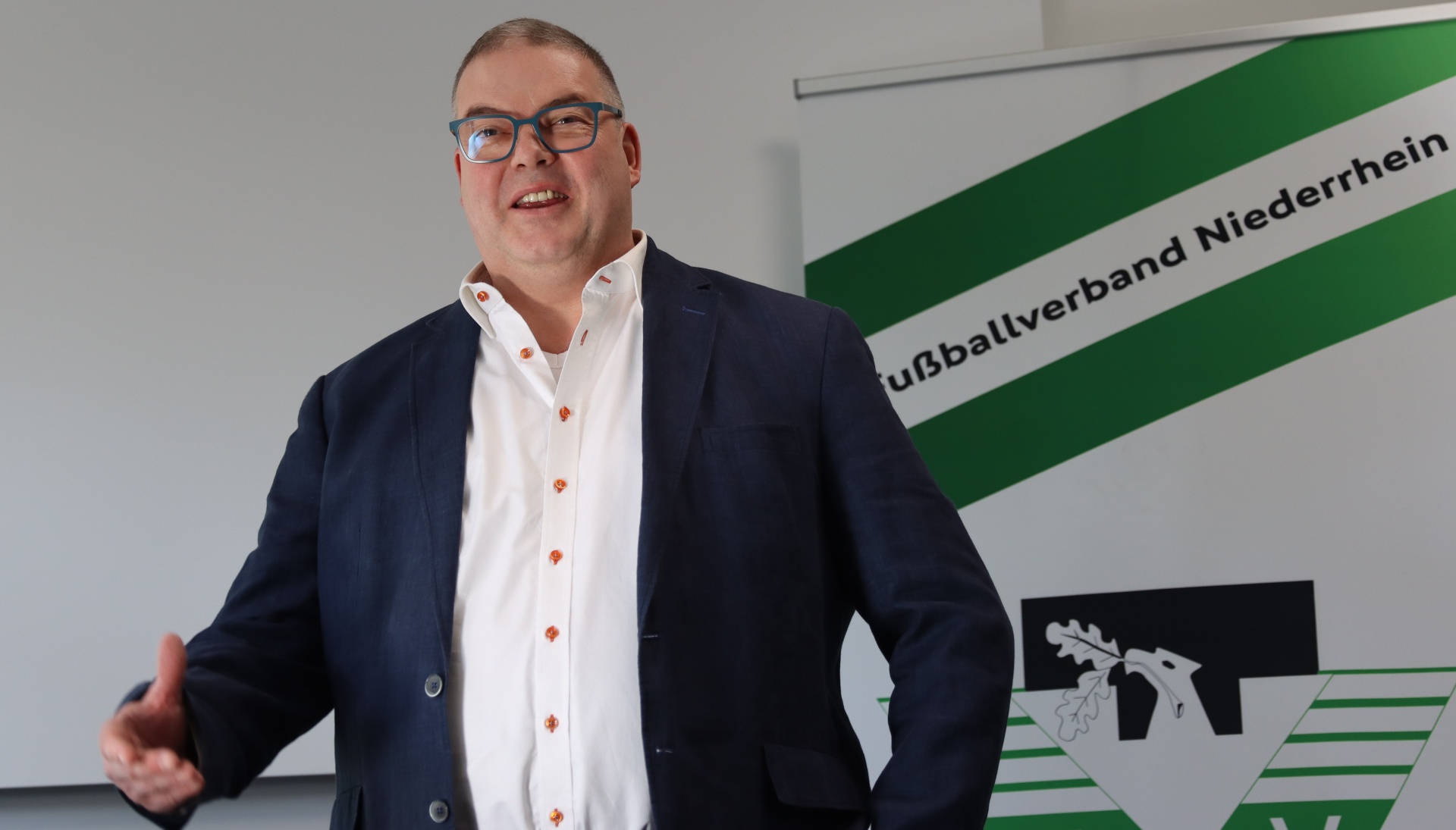[{"clang_id":1,"value":"Mann in Anzug gestikuliert vor grünem Banner mit Schriftzug \"Fußballverband Niederrhein\" und Logo mit Eiche."}]