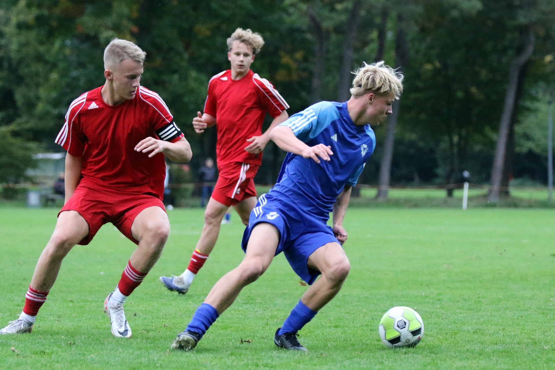 [{"clang_id":1,"value":"Drei Fußballspieler auf grünem Rasen. Ein Spieler in blau dribbelt den Ball, zwei in rot versuchen ihn zu blocken. Bäume im Hintergrund."}]