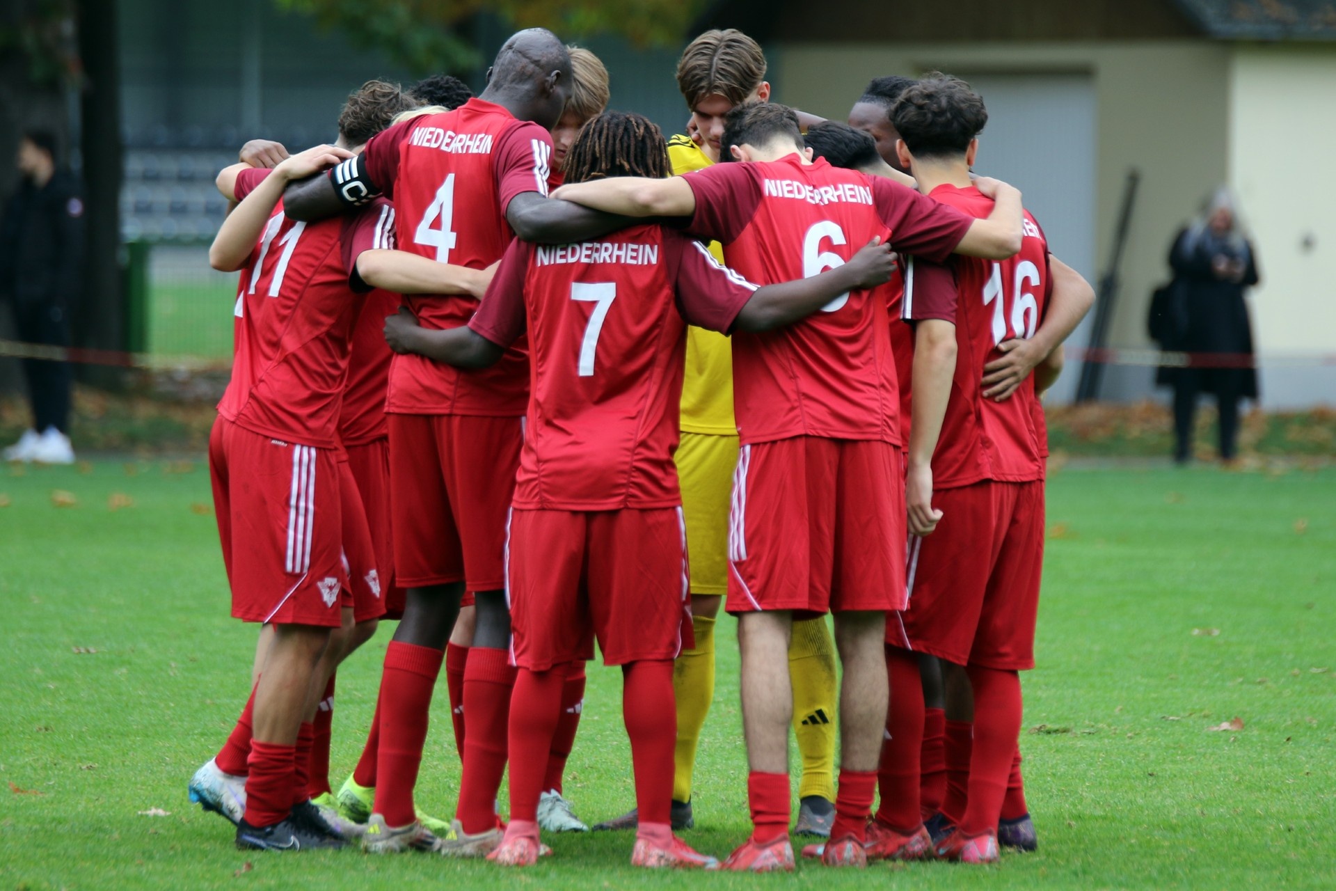 [{"clang_id":1,"value":"Fußballmannschaft in roten Trikots im Kreis stehend, Arme um die Schultern. Spieler tragen Nummern von 4 bis 16. Im Hintergrund Grünflächen und Zuschauer."}]