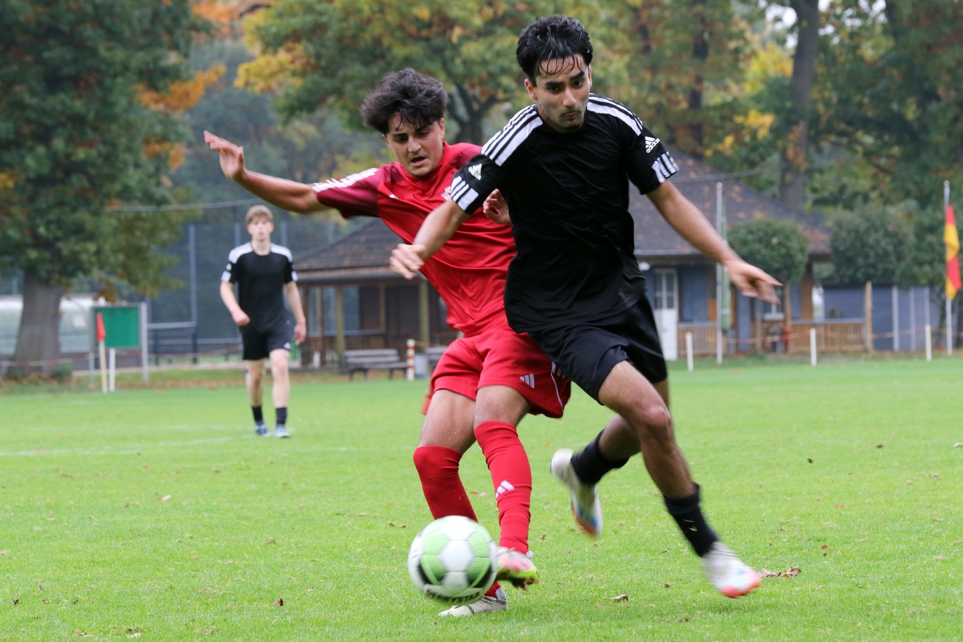 [{"clang_id":1,"value":"Zwei Fußballspieler im Zweikampf auf Grün. Ein Spieler trägt Rot, der andere Schwarz-Weiß. Im Hintergrund weitere Spieler und ein Gebäude."}]