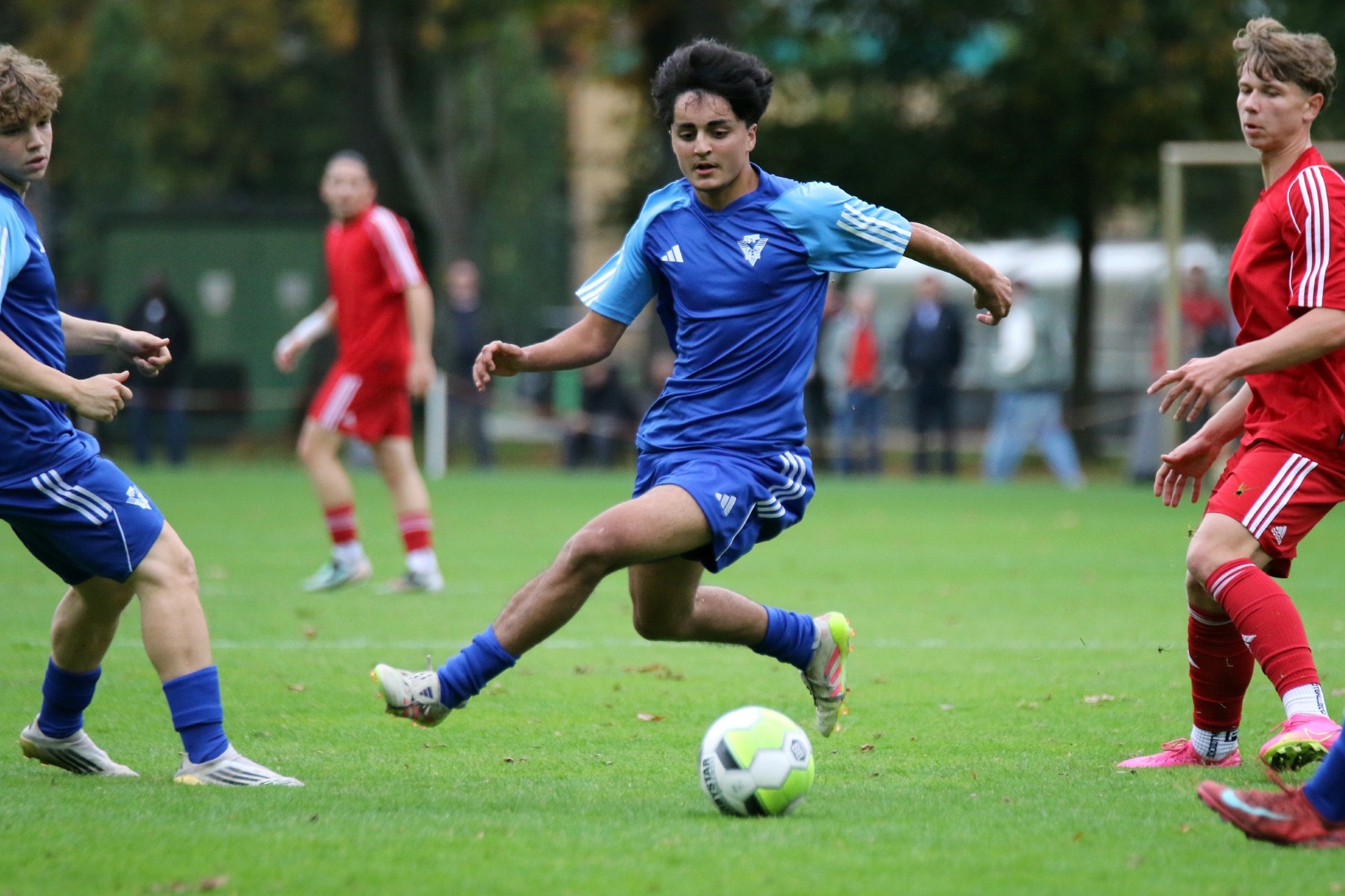 [{"clang_id":1,"value":"Fußballspiel: Jugendlicher in blauem Trikot schießt auf das Tor. Zwei weitere Spieler in Blau und Rot sind im Hintergrund zu sehen. Rasenplatz."}]