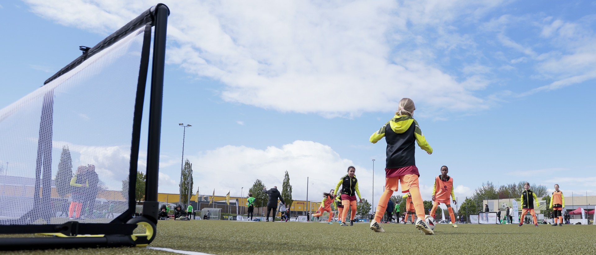 [{"clang_id":1,"value":"Fußballtraining: Spieler in orangefarbenen Trikots üben auf einem grünen Rasenfeld, ein Tor im Vordergrund. Himmel mit Wolken im Hintergrund."}]