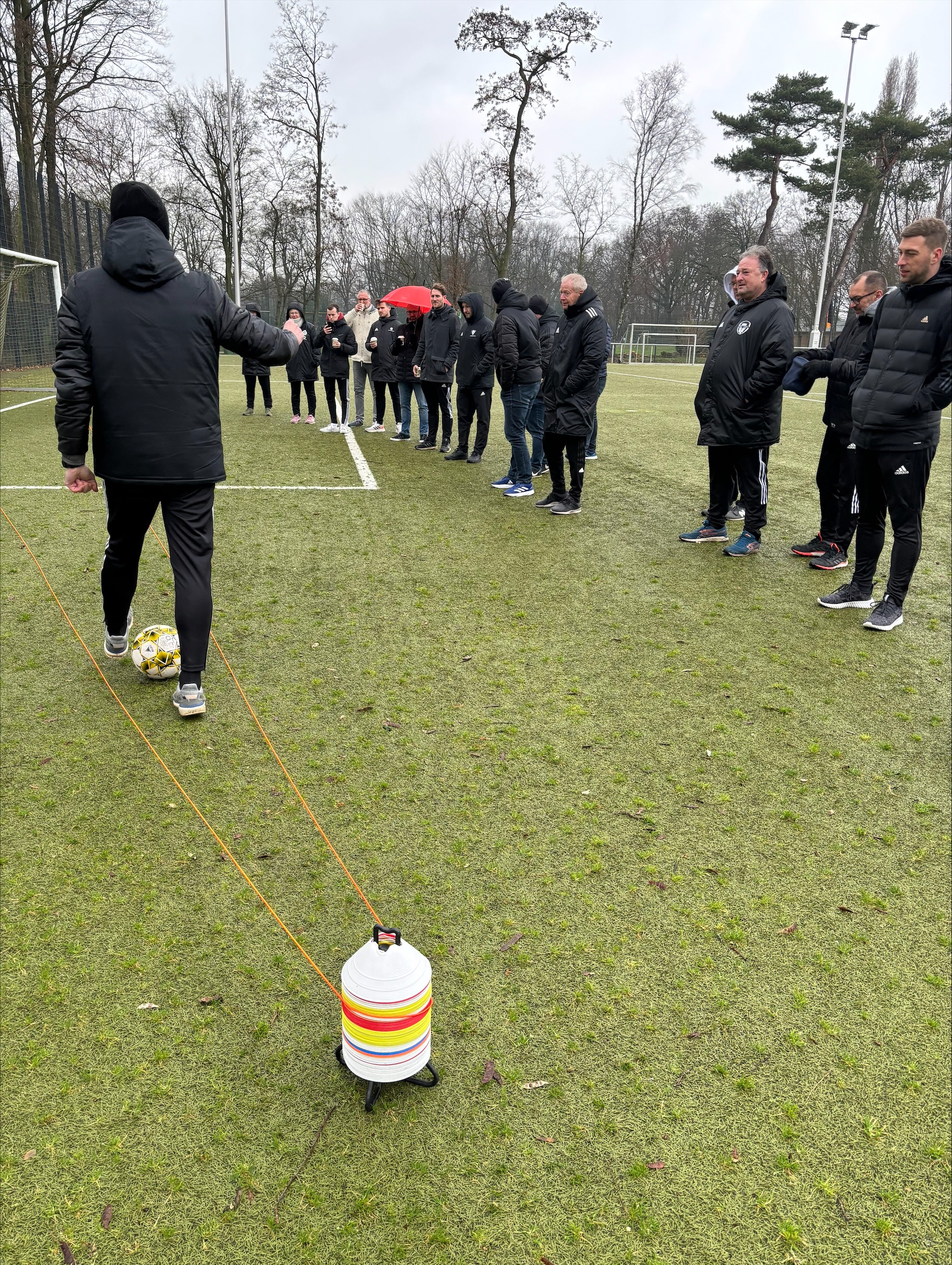 [{"clang_id":1,"value":"Fußballfeld mit Trainer und Gruppe von Männern. Eine Schnur spannt sich von einer Person zum Feldrand, markiert durch eine Wasserkanister-Konstruktion."}]