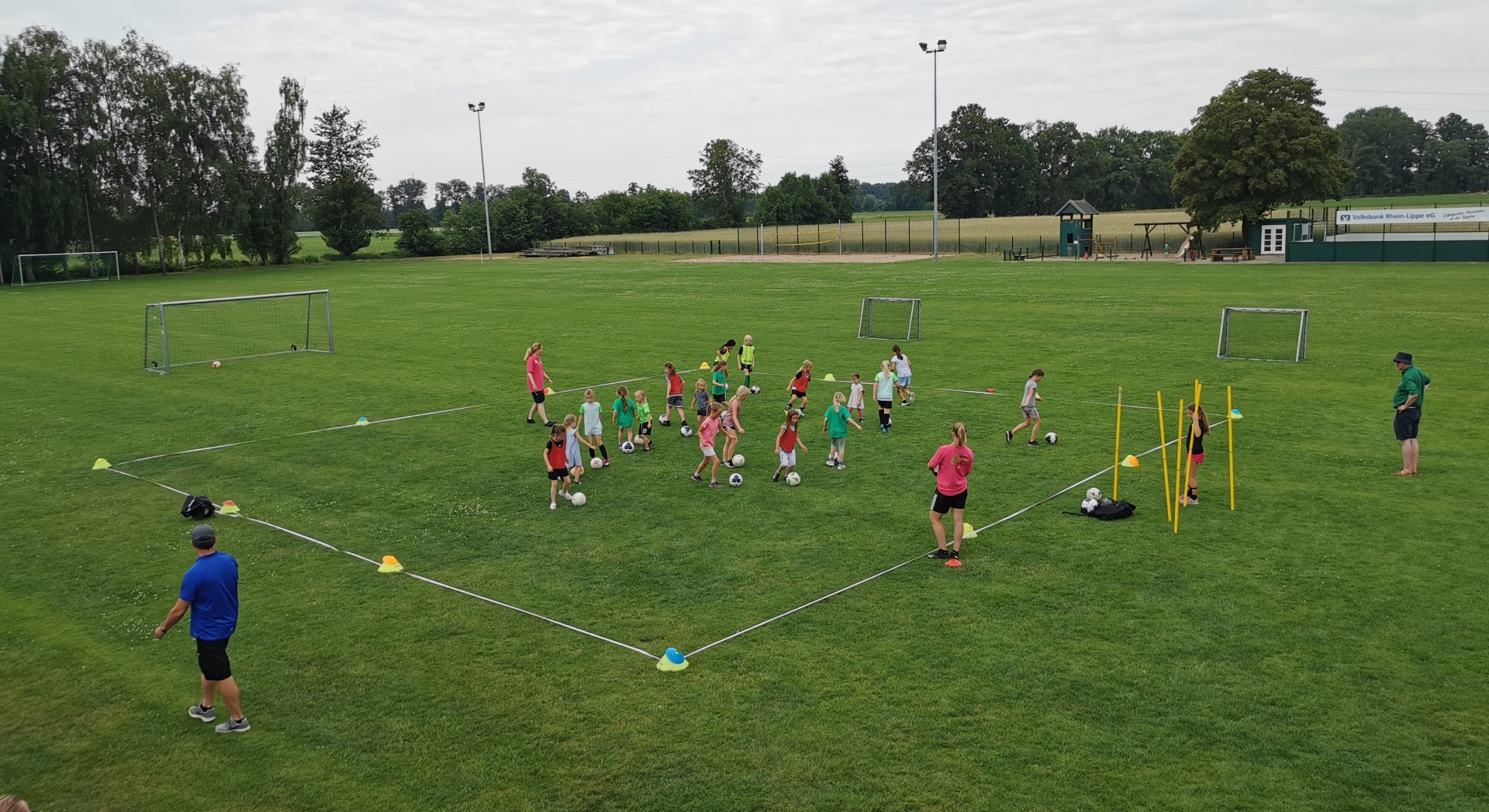 [{"clang_id":1,"value":"Fußballtraining mit Kindern auf Rasenplatz. Ein Trainer steht am Rand, Kinder in der Mitte, umgeben von Markierungshütchen. Im Hintergrund Bäume und ein Fußballtor."}]