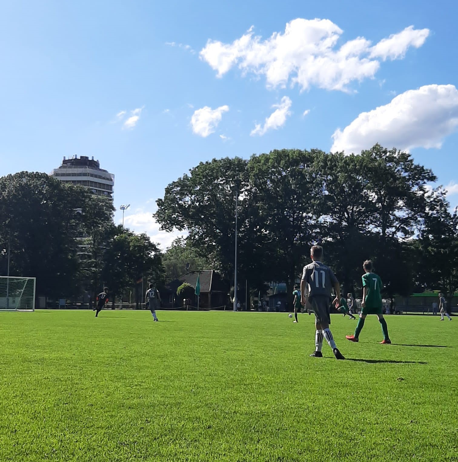 [{"clang_id":1,"value":"Fußballspiel im Freien. Spieler in grünen und weißen Trikots auf grünem Rasenfeld. Im Hintergrund Gebäude und Bäume unter blauem Himmel mit Wolken."}]