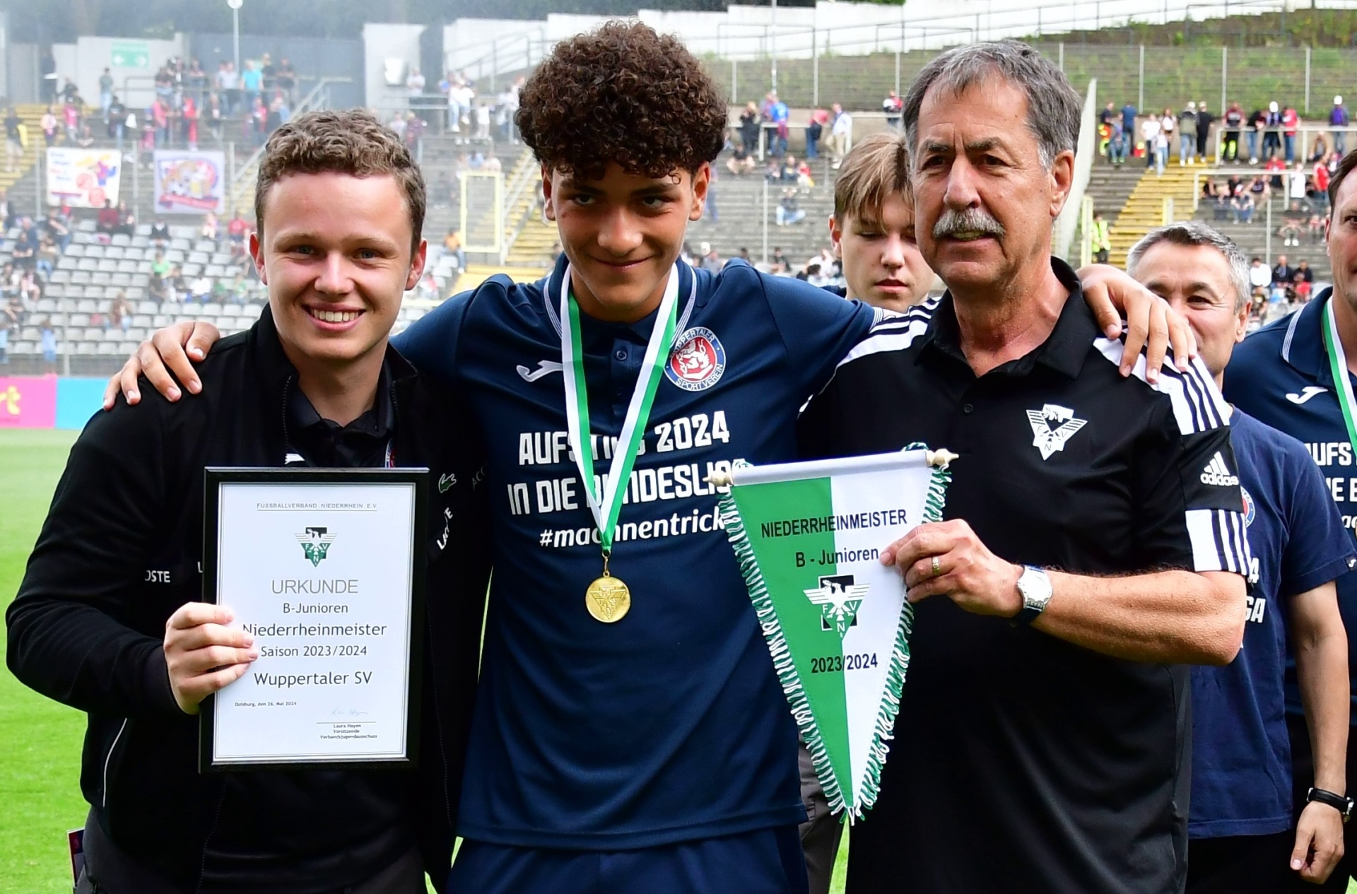 [{"clang_id":1,"value":"Fußballteam umrahmt Jugendlichen mit Auszeichnung \"B-Junioren Niederrheinmeister 2023/2024\". Im Hintergrund Fußballstadion und Zuschauer."}]