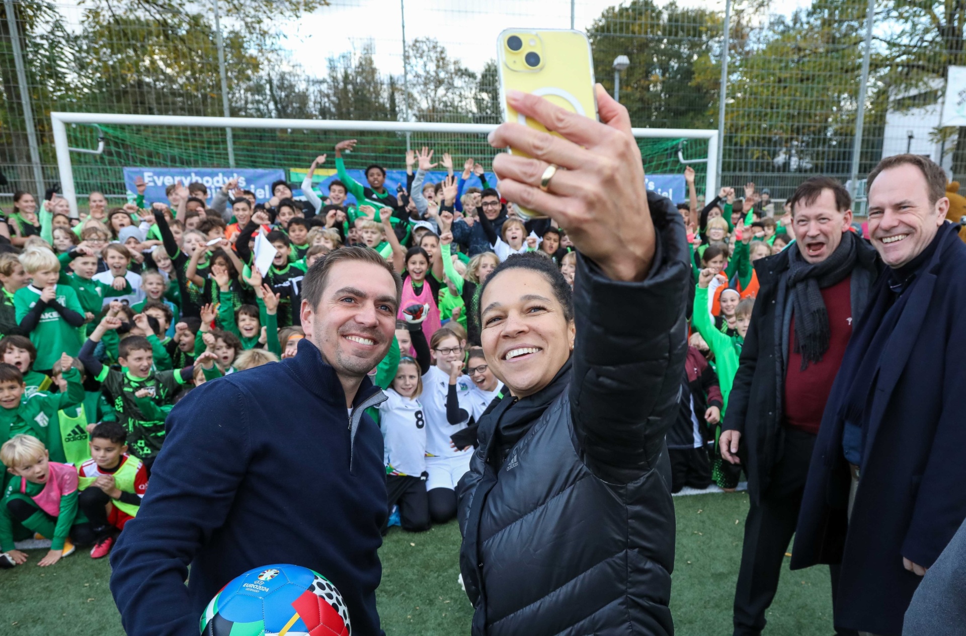 [{"clang_id":1,"value":"Gruppe von Menschen, darunter viele Kinder in Sportkleidung, posieren mit zwei Erwachsenen für ein Selfie auf einem Fußballfeld. Ein Fußball liegt im Vordergrund."}]