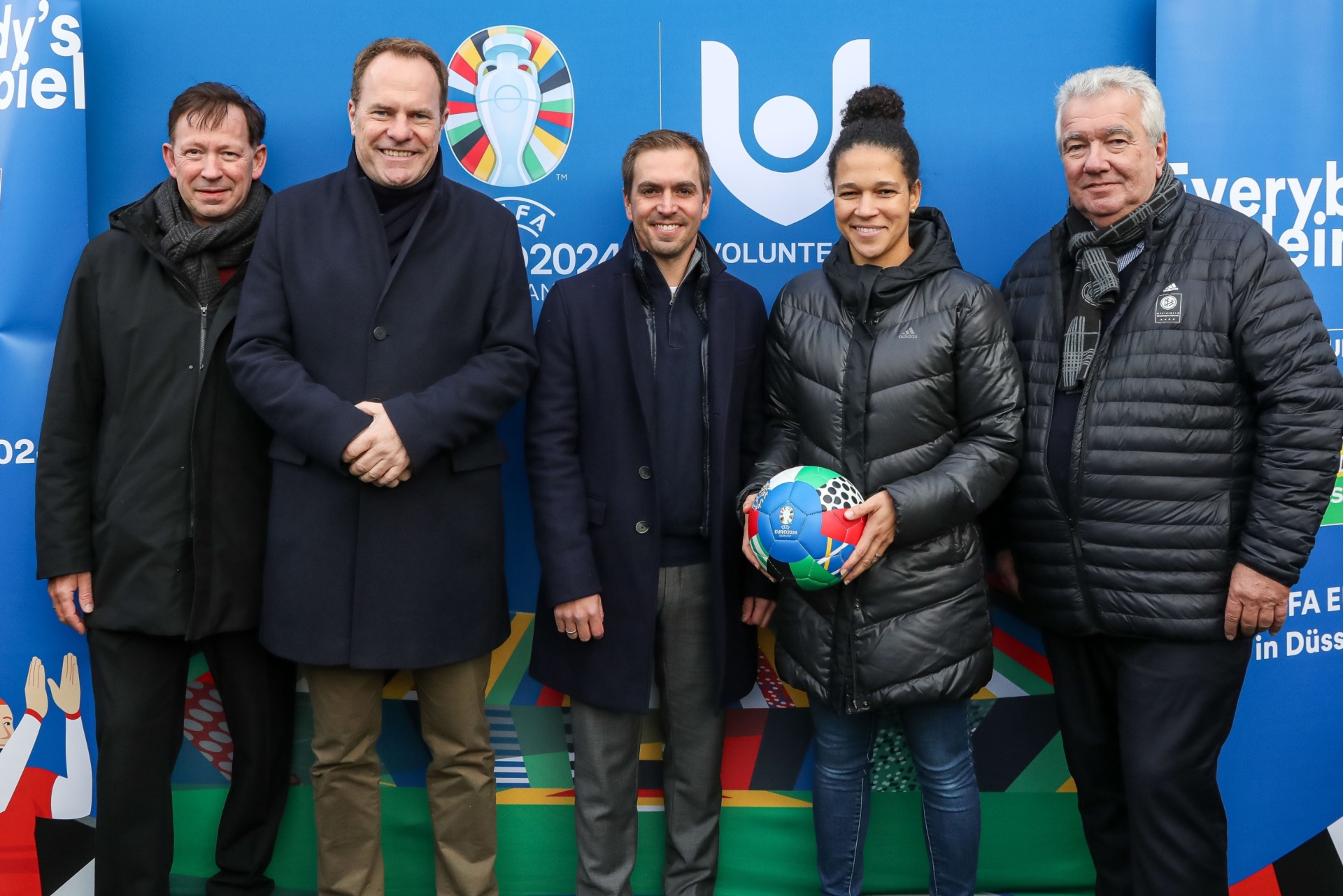 [{"clang_id":1,"value":"Fünf Männer und eine Frau stehen vor blauem Hintergrund mit Logo und Fahnen. Die Frau hält einen Fußball. Alle tragen Winterkleidung."}]