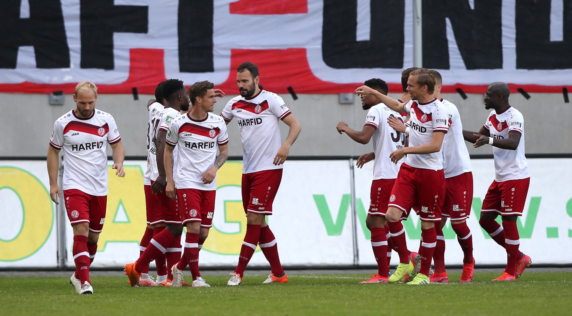 [{"clang_id":1,"value":"Fußballmannschaft in weißen Trikots mit roten Akzenten und Shorts auf grünem Rasen. Im Hintergrund Banner mit Schriftzügen und Zuschauerränge."}]