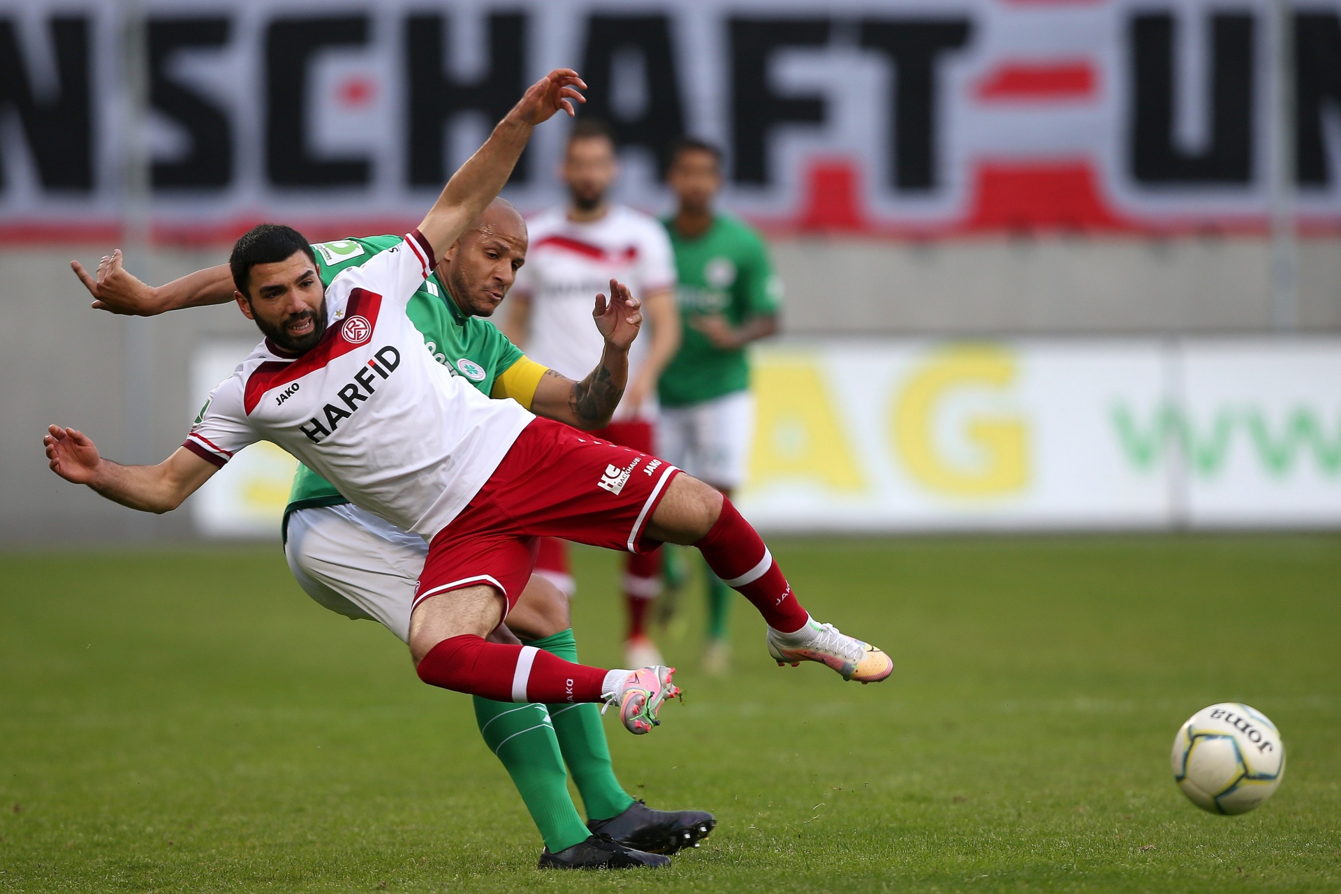 [{"clang_id":1,"value":"Zwei Fußballspieler kämpfen um den Ball auf grünem Rasen. Links ein Spieler in rot-weißem Trikot, rechts ein Spieler in grün-schwarzem Trikot. Im Hintergrund eine Werbetafel."}]