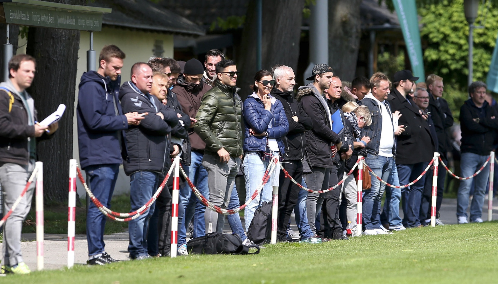 [{"clang_id":1,"value":"Gruppe Männer steht an einer Absperrung auf einer Wiese. Im Hintergrund Bäume und Gebäude."}]