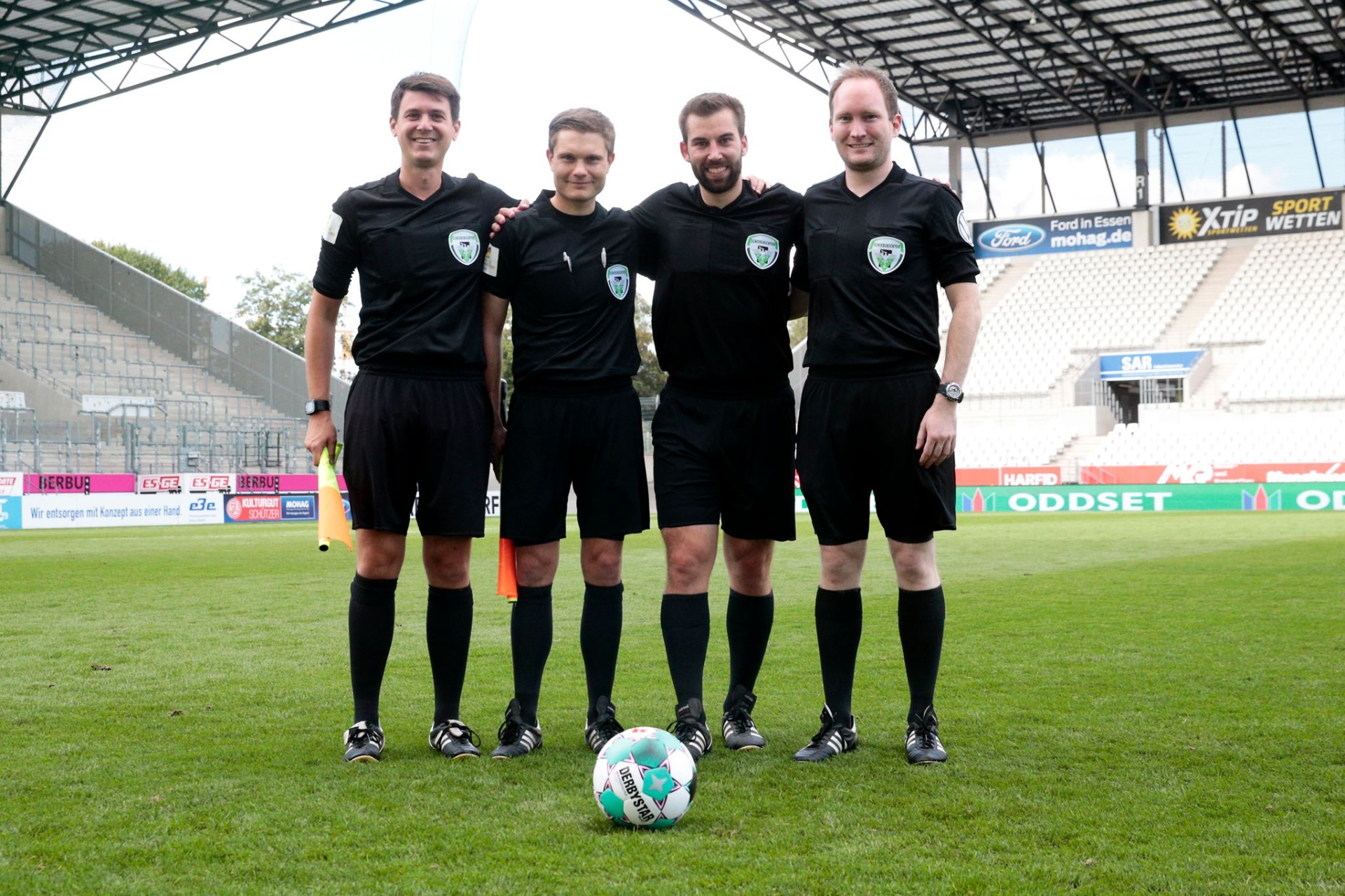 [{"clang_id":1,"value":"Vier Männer in schwarzer Fußballausstattung stehen auf einem Fußballfeld vor einem Stadion. Ein Fußball liegt vor ihnen."}]