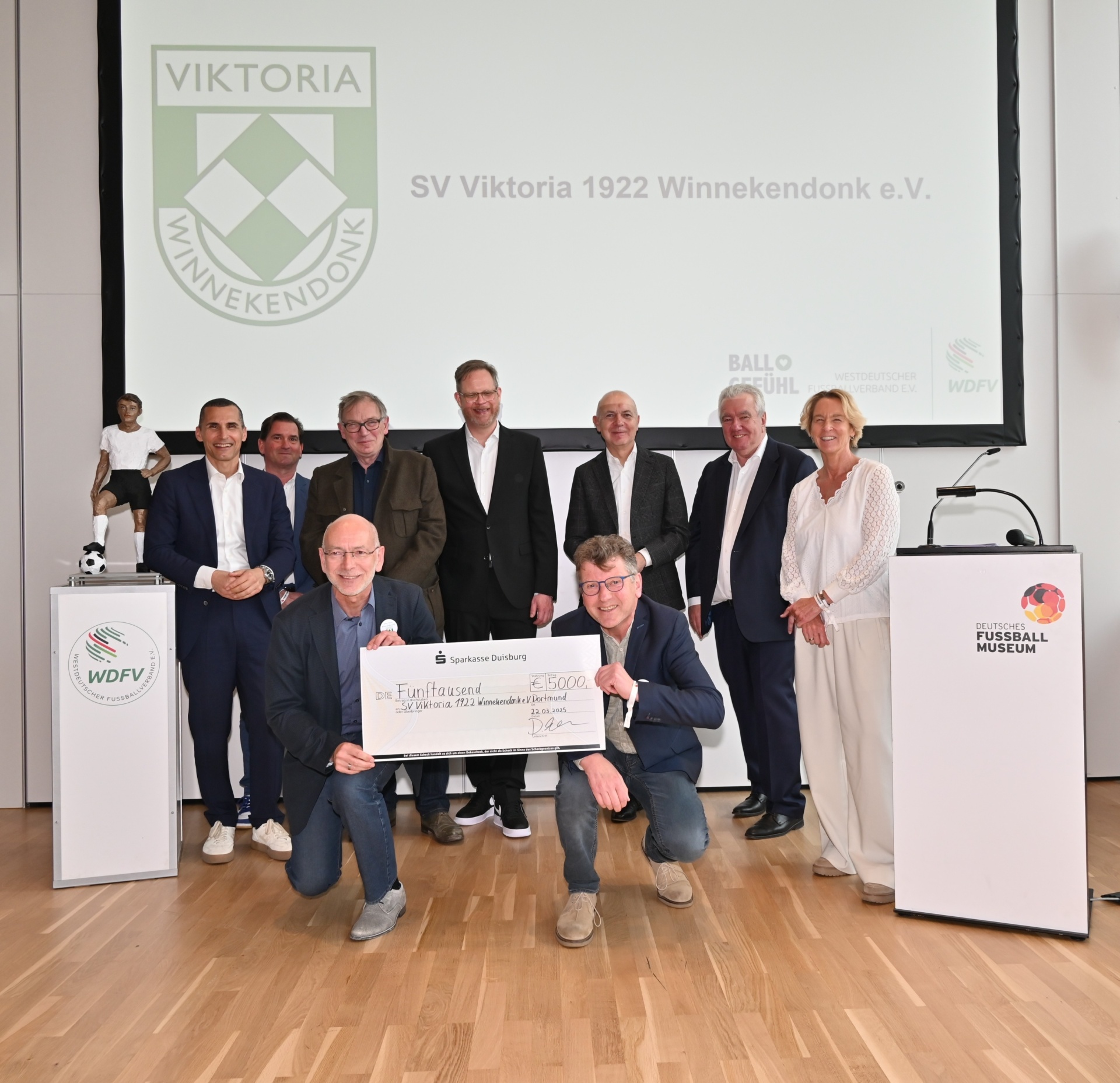 [{"clang_id":1,"value":"Gruppe posiert mit übergroßem Scheck (5000€) vor Vereinslogo \"Viktoria Winnekond\". Männer und Frauen in formeller Kleidung, teilweise kniend. Im Hintergrund Banner mit Vereinsnamen und Logos."}]