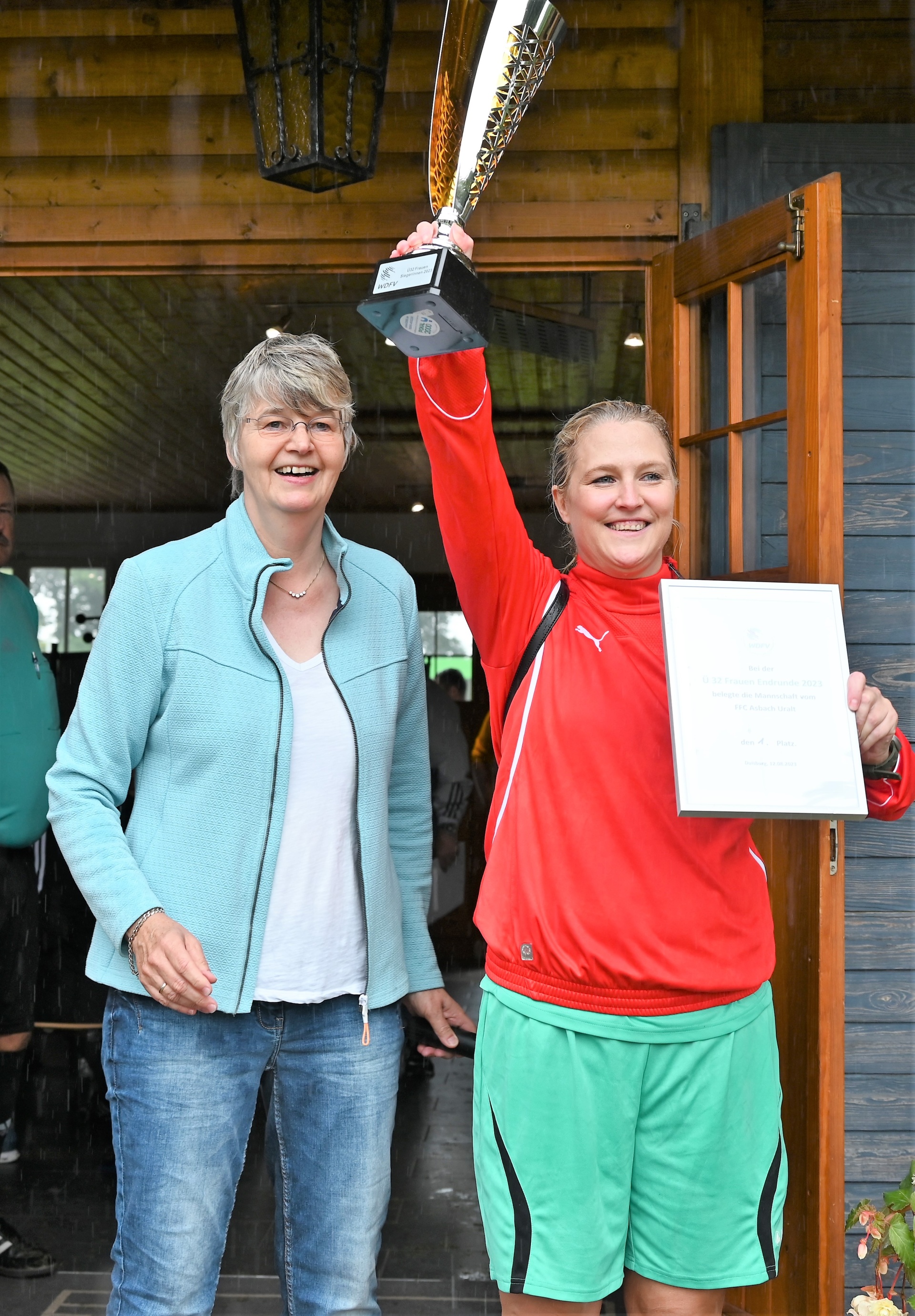 [{"clang_id":1,"value":"Zwei lächelnde Frauen posieren vor einer Holztür. Die Frau rechts hält einen Pokal und eine Urkunde hoch, trägt rote Sportkleidung und grüne Shorts. Die linke Frau trägt Jeans und einen blauen Pullover."}]