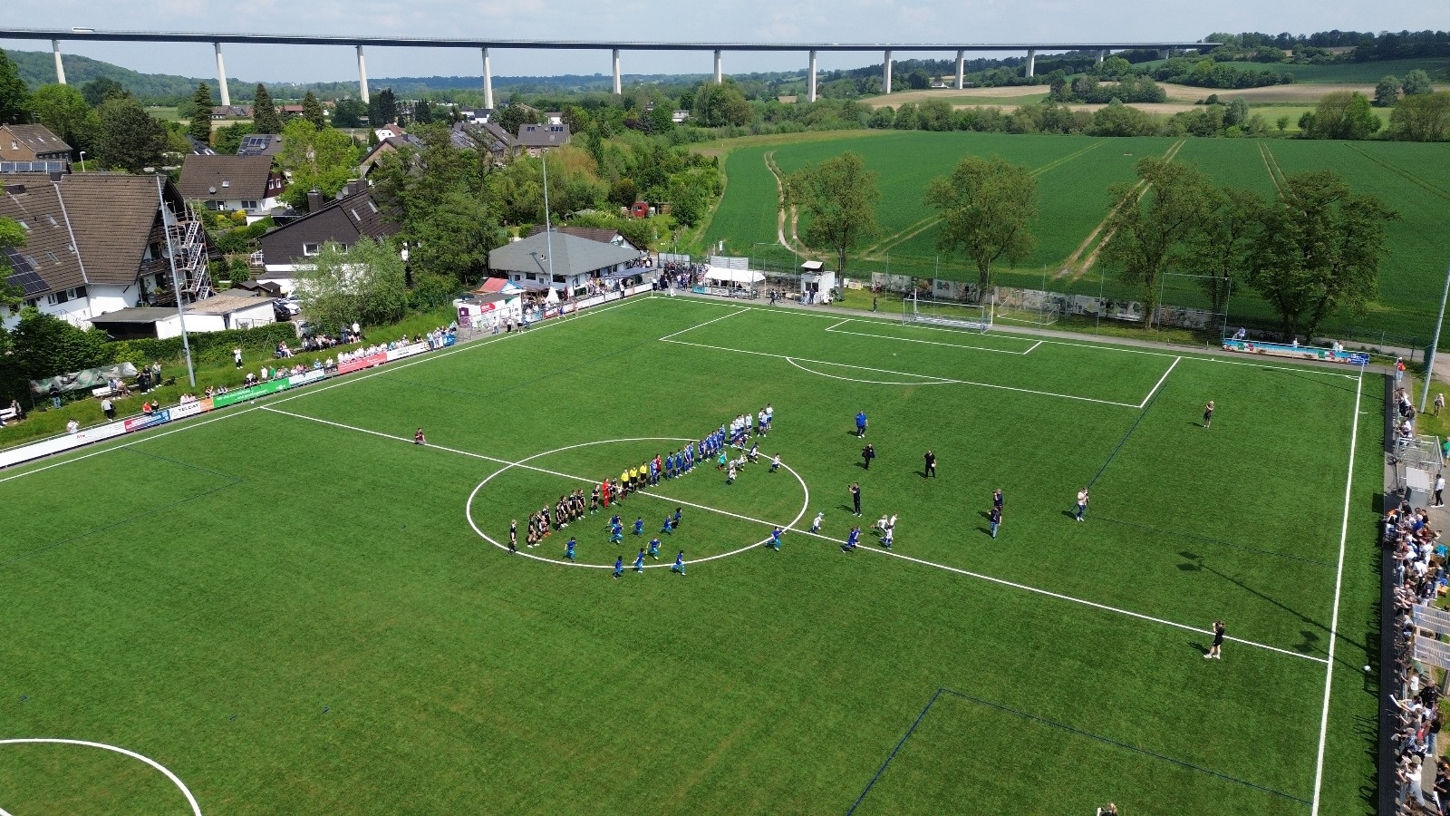 [{"clang_id":1,"value":"Fußballspiel auf grünem Rasenplatz. Zwei Mannschaften in blau und weiß kämpfen um den Ball. Im Hintergrund Häuser, Bäume und eine Brücke."}]