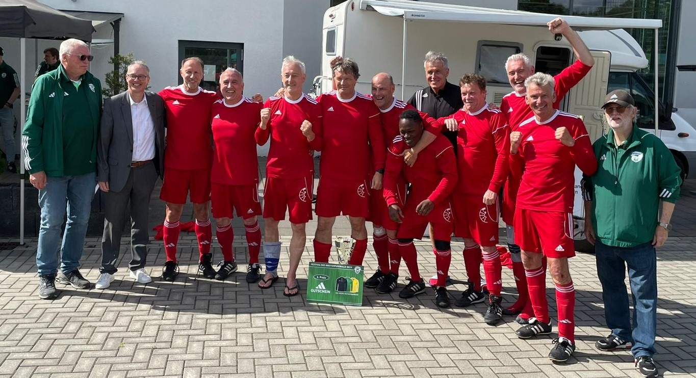 [{"clang_id":1,"value":"Fußballmannschaft in roten Trikots mit Pokal posiert vor Gebäuden und Wohnmobilen. Einige Personen tragen grüne Jacken."}]