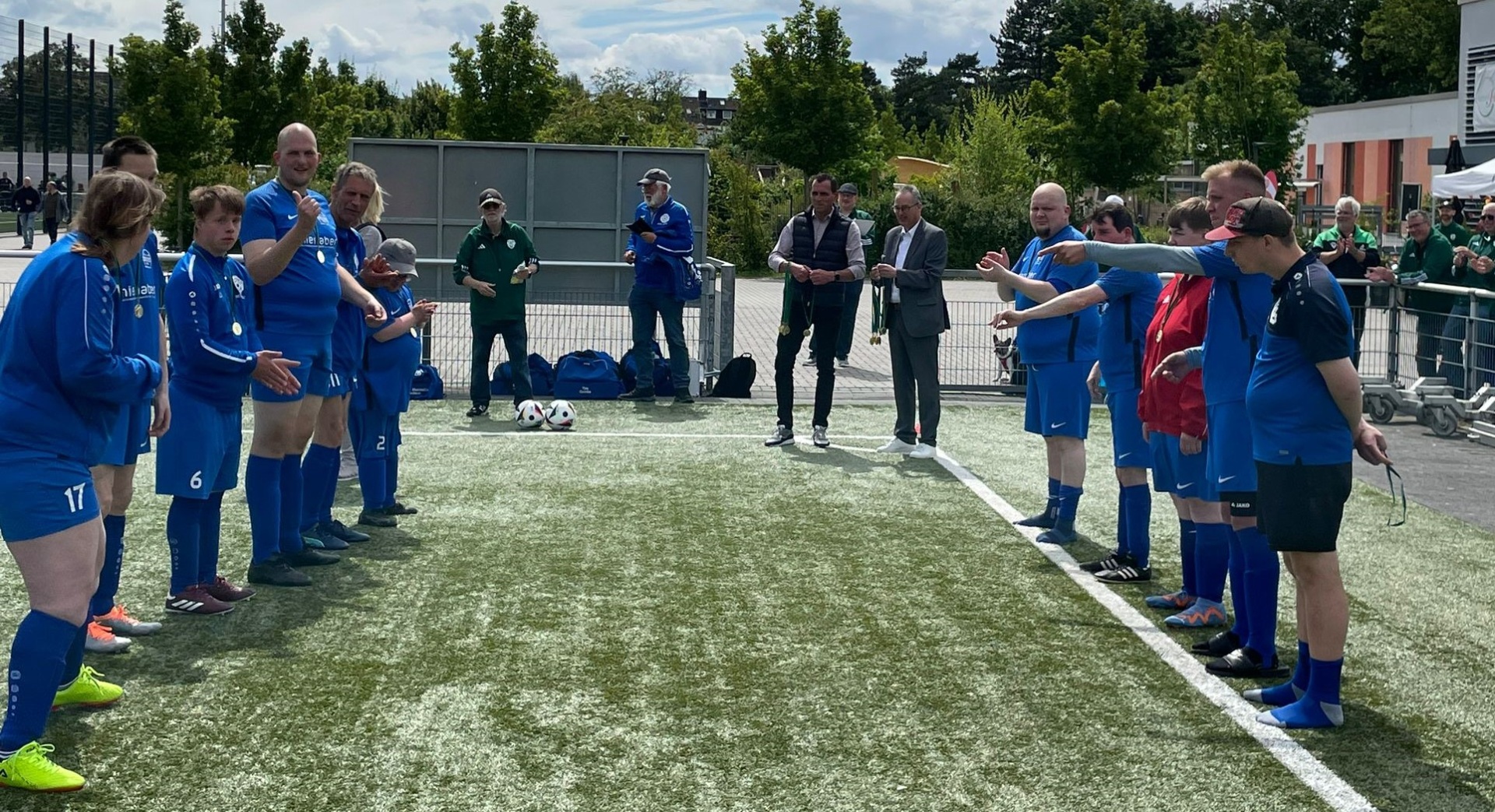 [{"clang_id":1,"value":"Fußballteam in blauen Trikots steht im Kreis auf einem Kunstrasenplatz. Einige Personen stehen außerhalb des Kreises, wahrscheinlich Trainer. Bäume im Hintergrund."}]