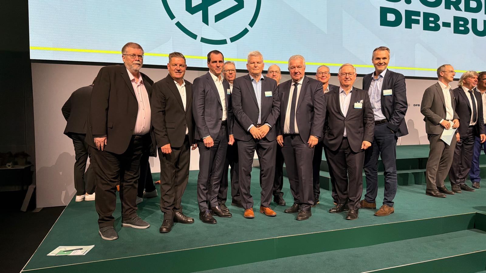[{"clang_id":1,"value":"Gruppe von etwa zwölf Männern in Anzügen auf einer grünen Bühne vor einem Hintergrund mit Logo und Text."}]