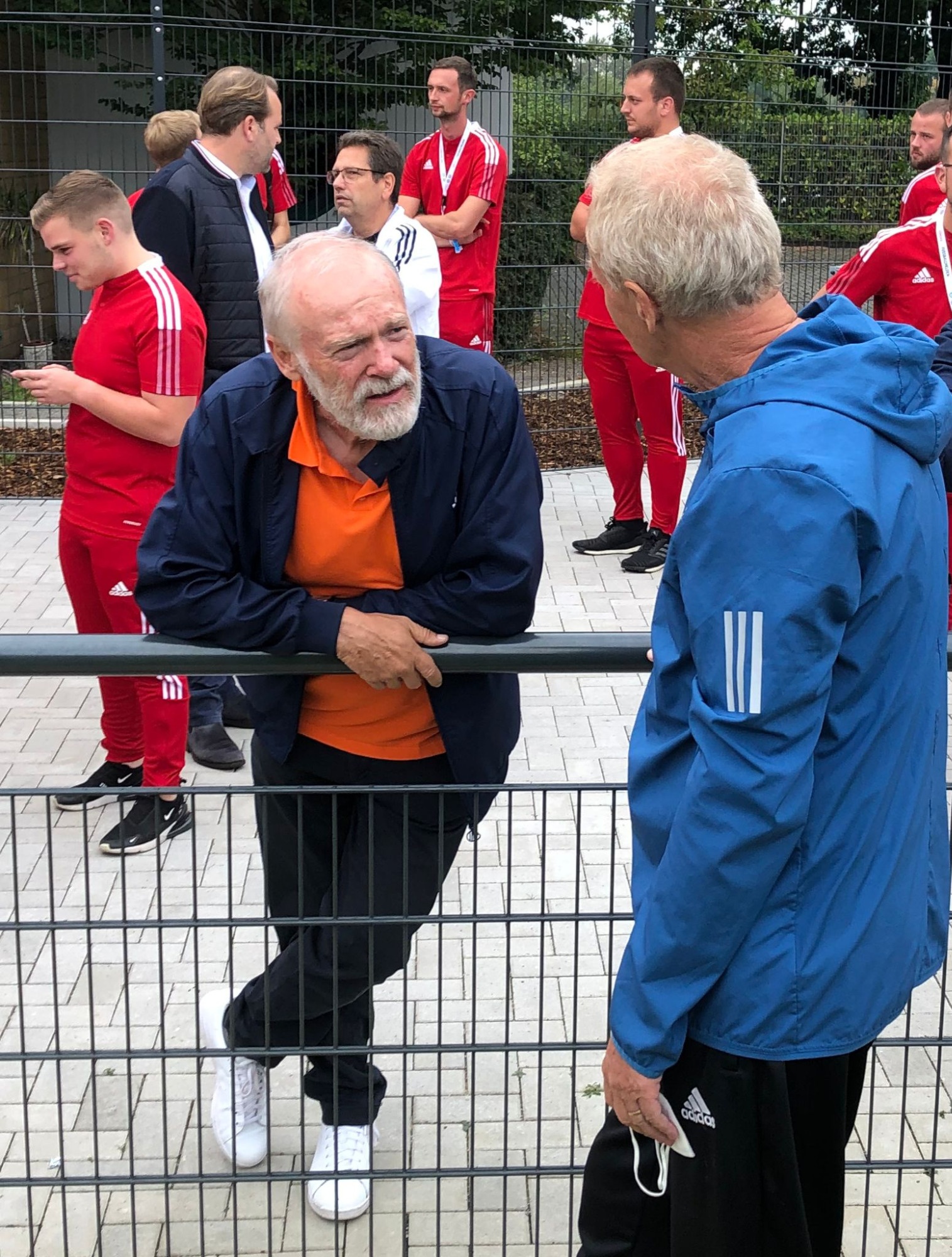 [{"clang_id":1,"value":"Zwei ältere Männer stehen an einem metallenen Zaun und sprechen. Im Hintergrund stehen Männer in roten Sportanzügen, vermutlich eine Fußballmannschaft."}]