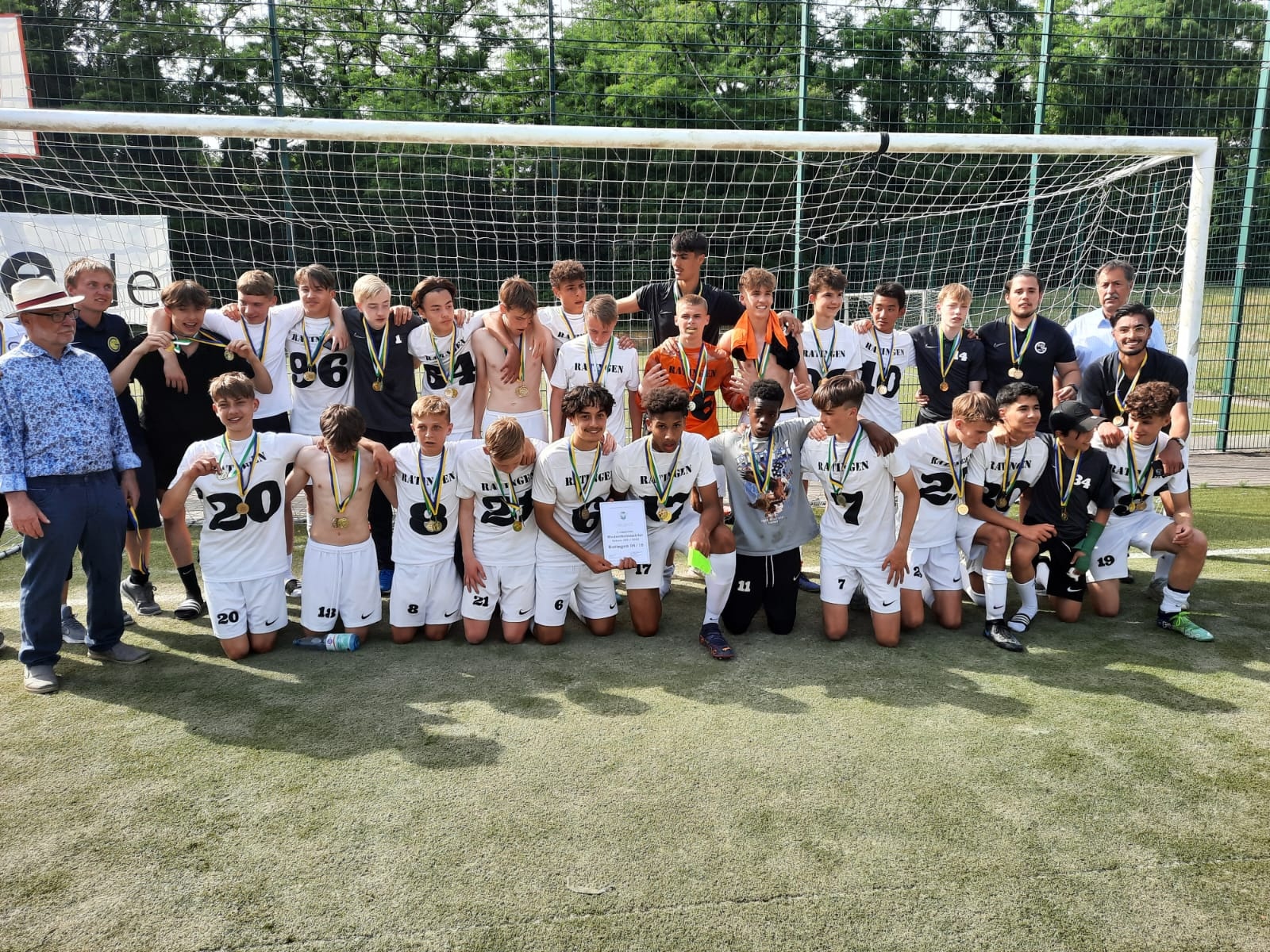 [{"clang_id":1,"value":"Fußballmannschaft mit Trainern und Betreuern, knien und stehen vor einem Fußballtor. Alle tragen Medaillen. Im Hintergrund Bäume und ein Zaun."}]
