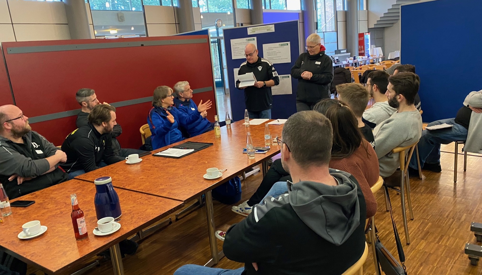 [{"clang_id":1,"value":"Gruppe diskutiert an einem grossen Tisch in einer Halle. Einige Personen sitzen, andere stehen. Im Hintergrund weitere Personen und Poster."}]
