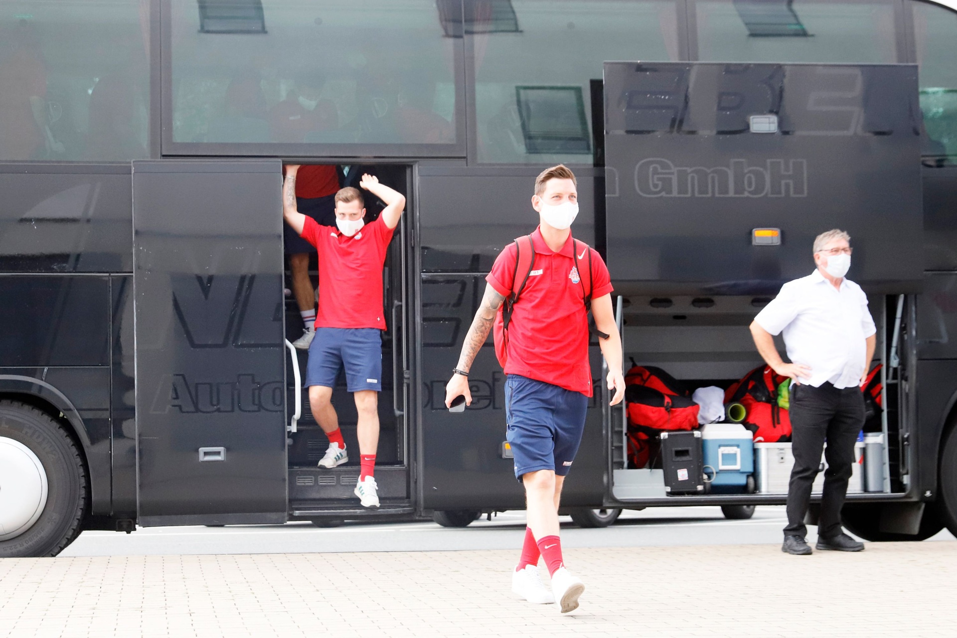 [{"clang_id":1,"value":"Zwei Fußballspieler in roten Trikots und Shorts steigen aus einem dunklen Mannschaftsbus. Beide tragen Masken. Im Hintergrund eine Person mit Hemd und Hose."}]