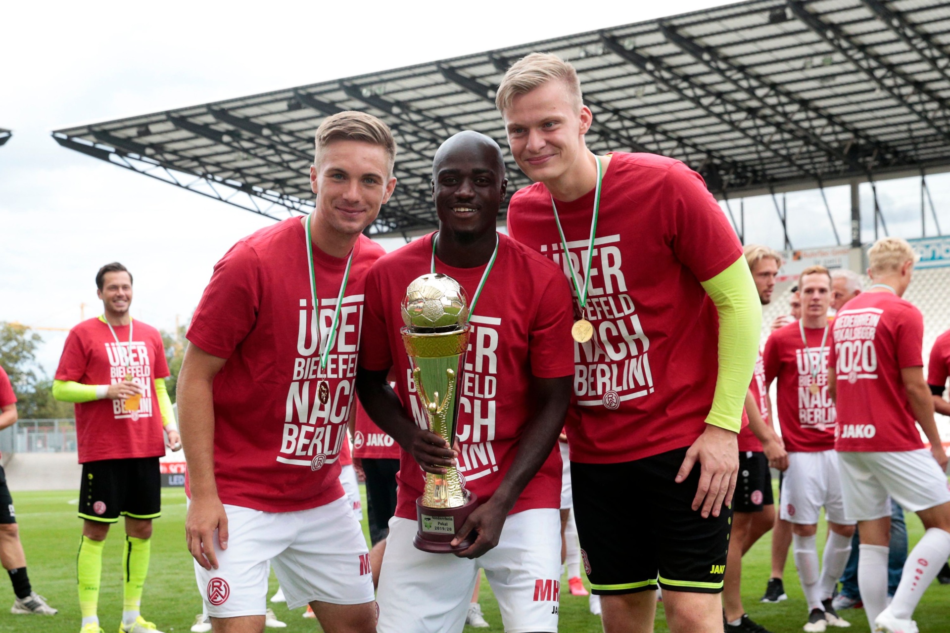 [{"clang_id":1,"value":"Drei Fußballspieler mit Pokal. Im Hintergrund weitere Spieler und ein Stadiongebäude. Alle tragen rote T-Shirts mit Aufdruck und weiße Shorts."}]