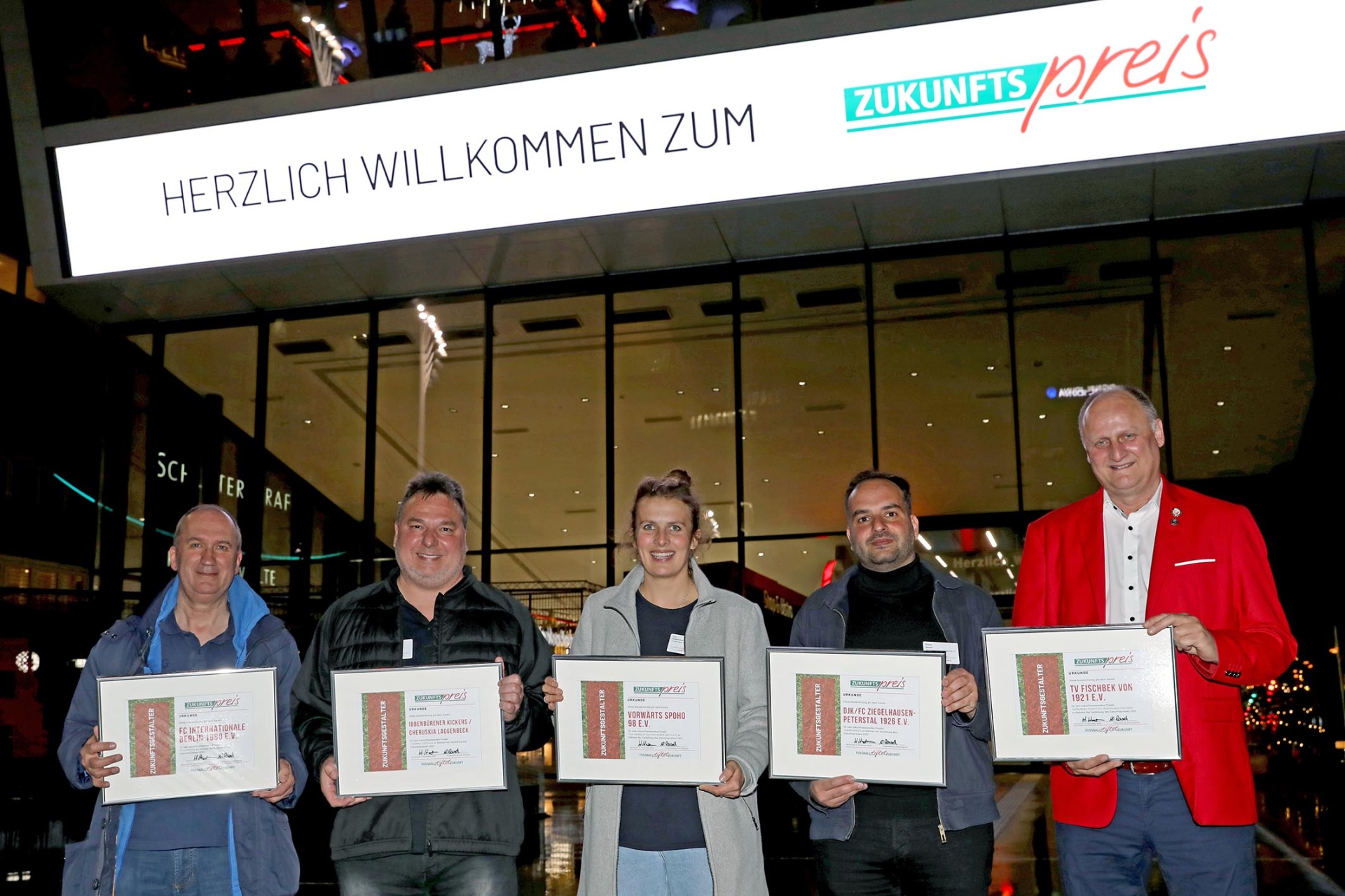 [{"clang_id":1,"value":"Fünf Personen stehen vor einer verglasten Fassade mit dem Schriftzug \"HERZLICH WILLKOMMEN ZUM ZUKUNFTSpreis\" und halten Urkunden."}]