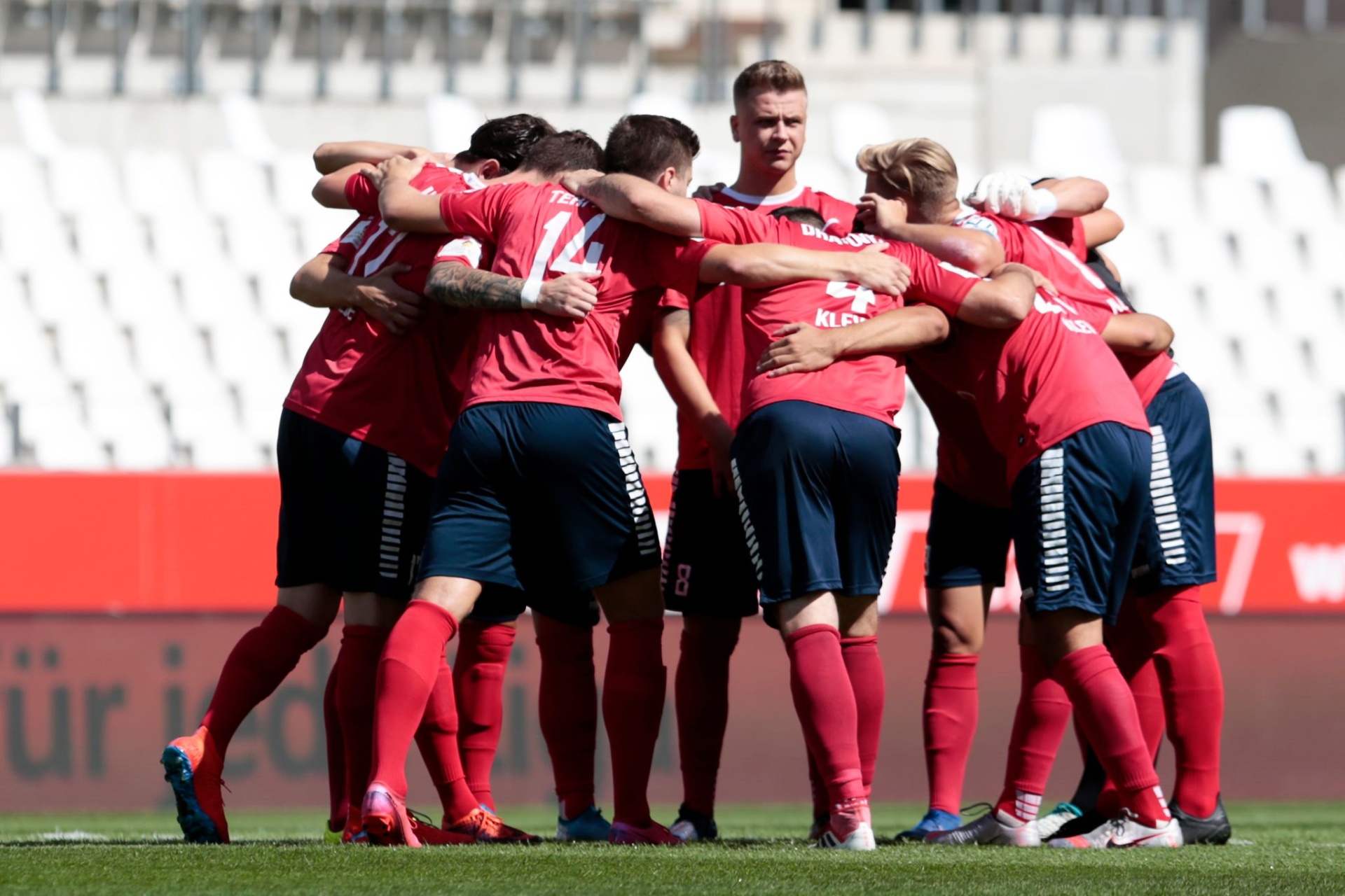 [{"clang_id":1,"value":"Fußballspieler im roten Trikot und blauen Shorts feiern eng umschlungen auf grünem Rasen. Im Hintergrund ein Stadion."}]