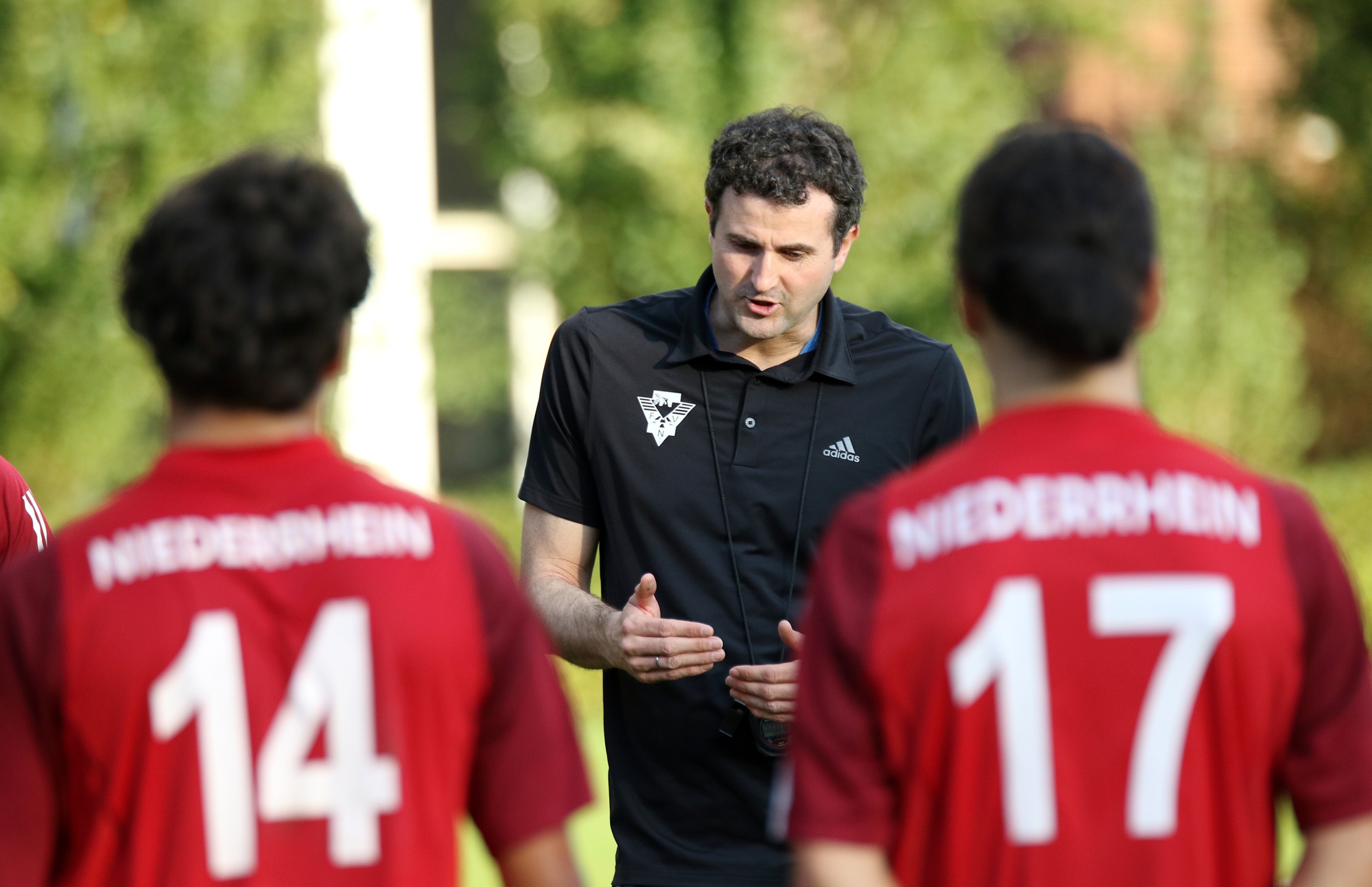 Fußballtrainer mit Whistle spricht zwei Spieler in roten Trikots mit den Nummern 14 und 17 auf einem Fußballfeld. Im Hintergrund verschwommene Bäume und ein Gebäude.