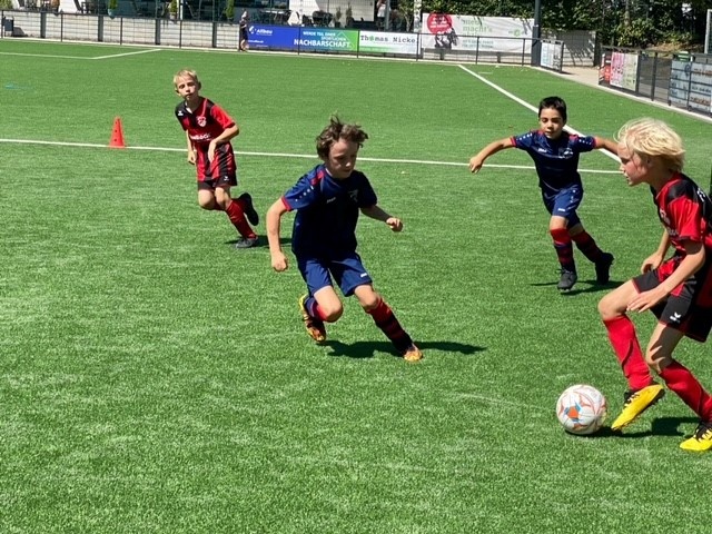 [{"clang_id":1,"value":"Vier Kinder spielen Fußball auf einem grünen Kunstrasenplatz. Zwei tragen rote Trikots, zwei blaue. Ein Kind schießt auf den Ball. Im Hintergrund eine Absperrung und Werbebanner."}]