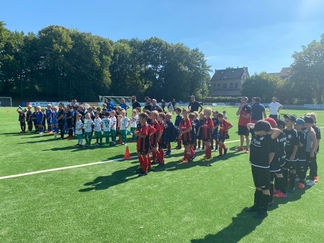 [{"clang_id":1,"value":"Fußballfeld mit zwei Jugendmannschaften, die sich zur Begrüßung gegenüberstehen. Im Hintergrund Gebäude und Bäume unter blauem Himmel."}]