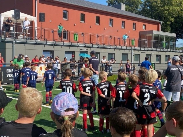 [{"clang_id":1,"value":"Kinderfußballspiel auf Rasenplatz. Zwei Mannschaften in roten und blauen Trikots stehen sich gegenüber. Zuschauer stehen am Spielfeldrand und auf einer Balkonterrasse eines Gebäudes im Hintergrund."}]