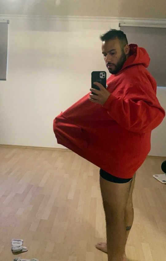 [{"clang_id":1,"value":"Mann mit rotem Hoodie, der diesen vorne aufhält. Er trägt dunkle Shorts und ist barfuß in einem hellen Raum mit Parkettboden."}]