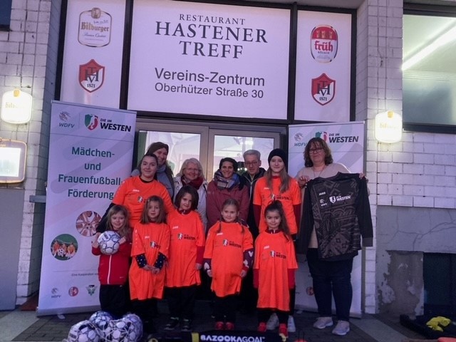 [{"clang_id":1,"value":"Gruppe von jungen Fußballspielerinnen in orangefarbenen Trainingsanzügen vor Restaurant \"Hastener Treff\", mit Bannern zur Förderung von Mädchen- und Frauenfußball."}]