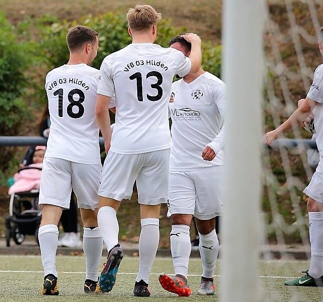 [{"clang_id":1,"value":"Fußballspieler mit weißen Trikots und den Rückennummern 13 und 18 stehen auf einem Fußballfeld. Im Hintergrund ein Zaun und Zuschauer."}]
