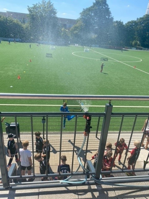 [{"clang_id":1,"value":"Fußballfeld mit spielenden Kindern und Sprinkleranlage. Im Vordergrund Sitzreihen mit weiteren Kindern, die zuschauen."}]