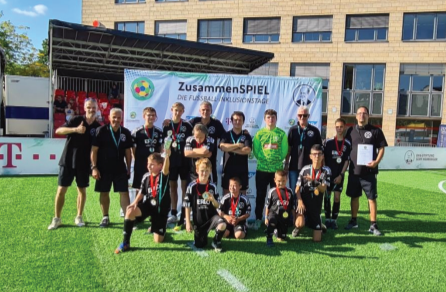 [{"clang_id":1,"value":"Fußballmannschaft mit Betreuern auf grünem Rasen vor einem Banner mit dem Schriftzug \"ZusammenSPIEL\". Alle tragen Sportkleidung, einige haben Medaillen um den Hals. Im Hintergrund Gebäude und ein T-Mobil-Logo."}]