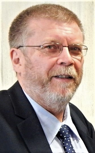 [{"clang_id":1,"value":"Mann mit grauem Bart und Brille, gekleidet in Anzug und Hemd mit Krawatte, lächelt in die Kamera. Heller Hintergrund."}]