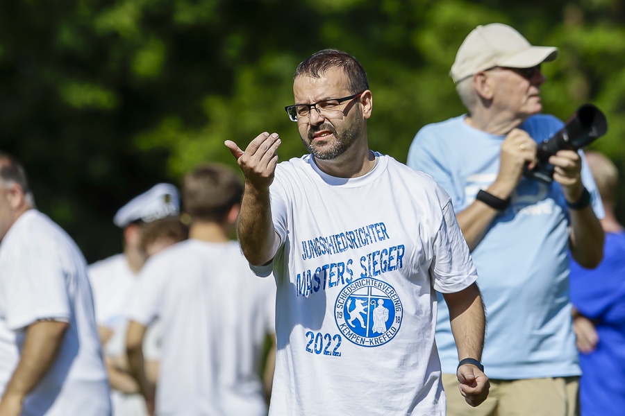 [{"clang_id":1,"value":"Mann mit T-Shirt \"Jungschiedsrichter Masters Sieger 2022\" gestikuliert im Freien, im Hintergrund verschwommene Personen und Bäume."}]