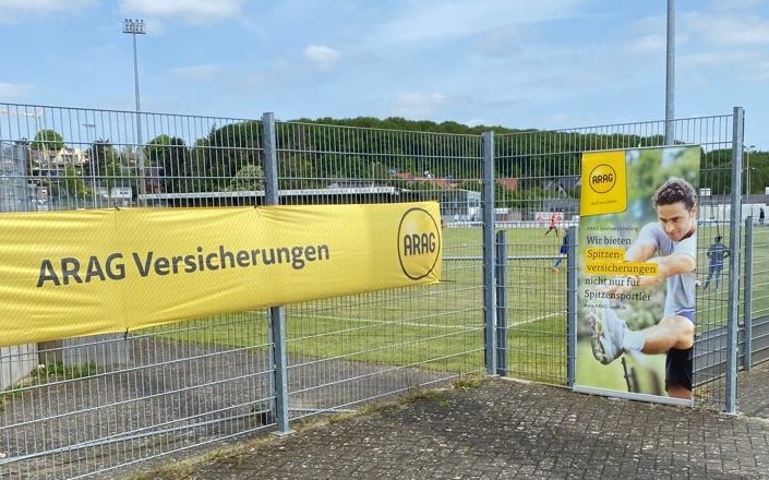 [{"clang_id":1,"value":"Werbebanner \"ARAG Versicherungen\" an einem Metallzaun eines Sportplatzes. Rechts ein Plakat mit Sportler und Text in deutscher Sprache. Hintergrund: Grünflächen und Gebäude."}]