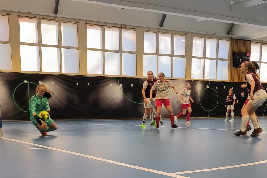 [{"clang_id":1,"value":"Futsal-Spiel in Sporthalle. Spielerinnen in rot und weiß kämpfen um den Ball. Im Hintergrund Fenster und Anzeigetafel."}]