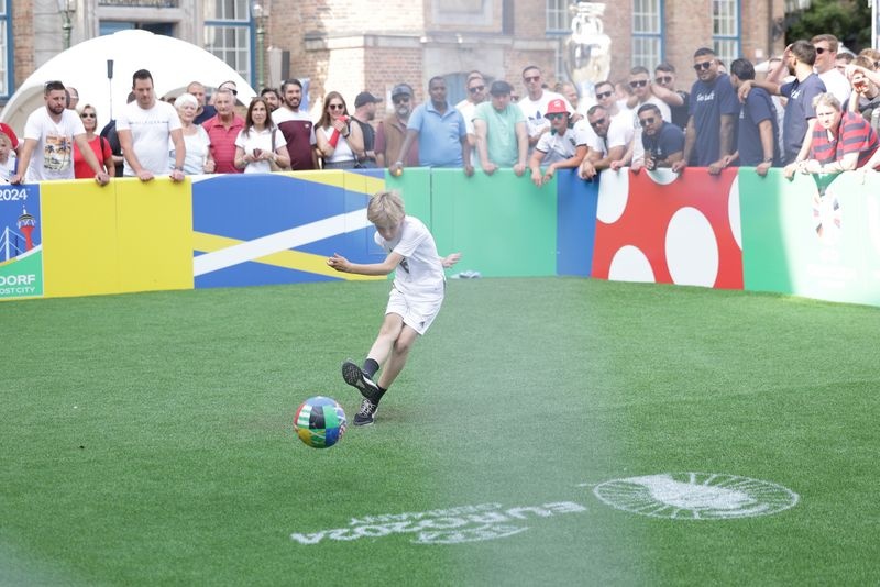 [{"clang_id":1,"value":"Junge kickt Fußball auf grünem Rasen vor Zuschauermenge. Im Hintergrund bunte Bande mit \"DORT CITY 2024\"-Schriftzug und Gebäude."}]